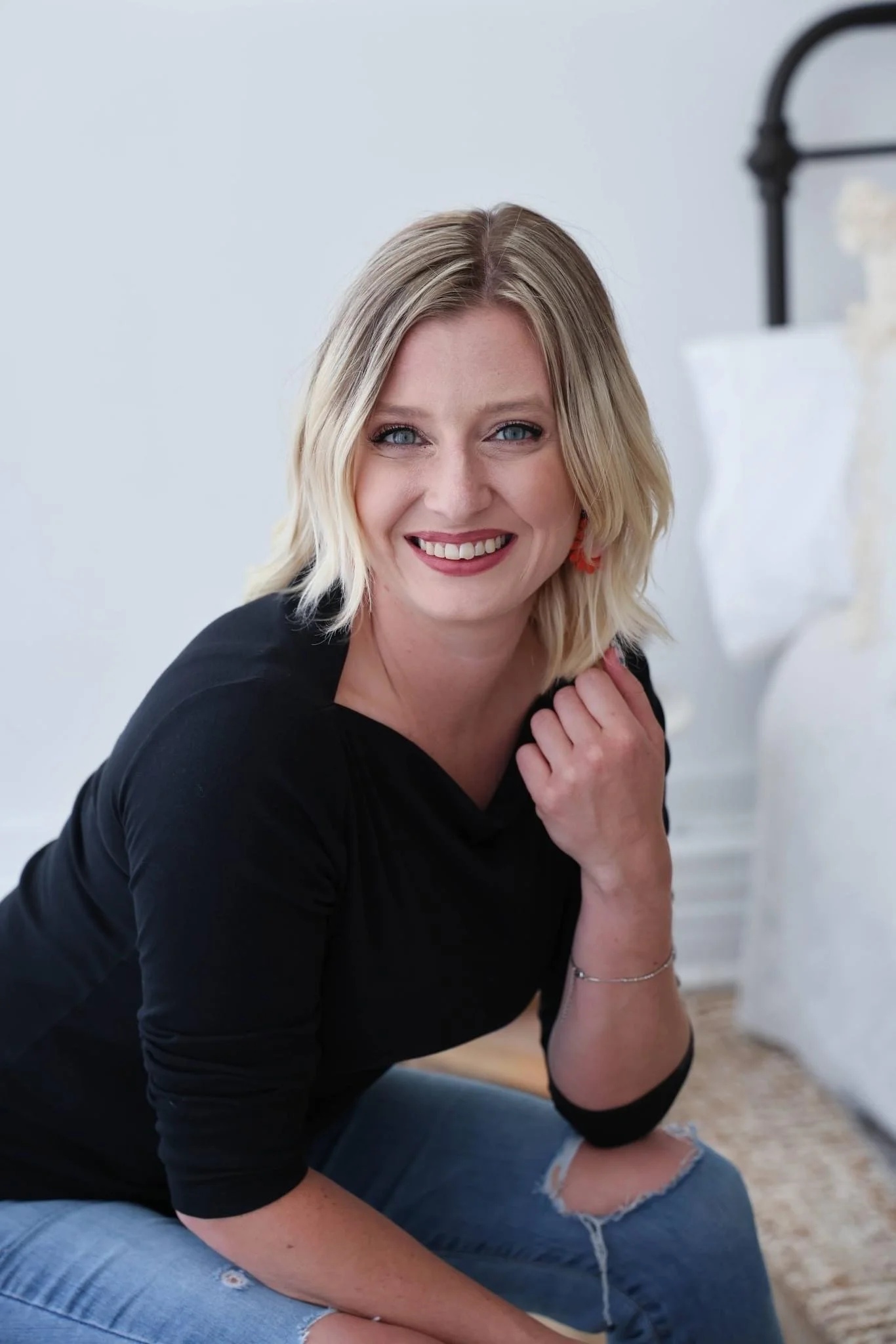 A woman with blonde hair and blue eyes smiling, wearing a black top and ripped jeans, sitting on the floor near a bed with white bedding.