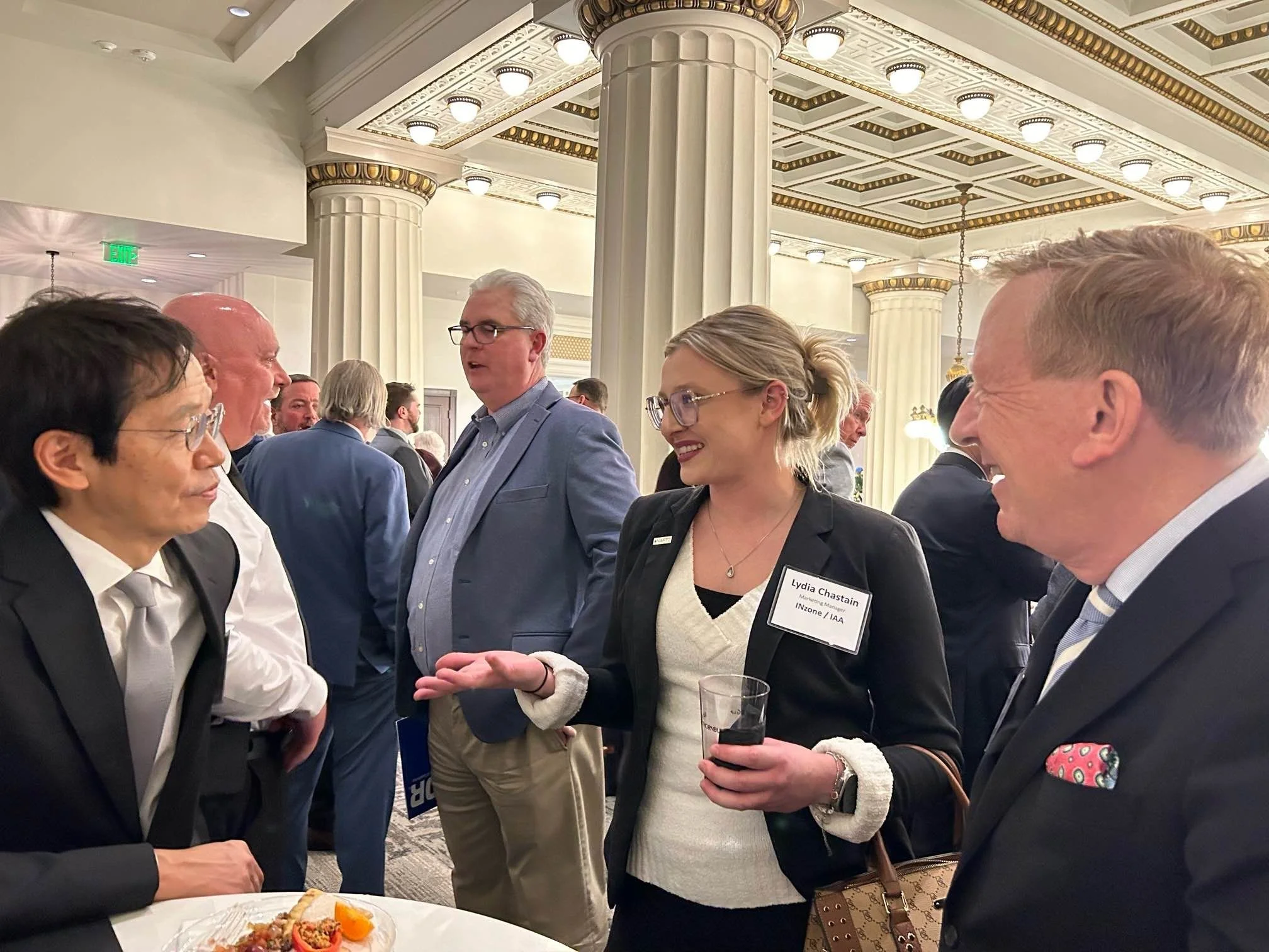 Four people engaged in conversation at a professional networking event, with a woman speaking and three men listening, in a decorated, elegant venue.