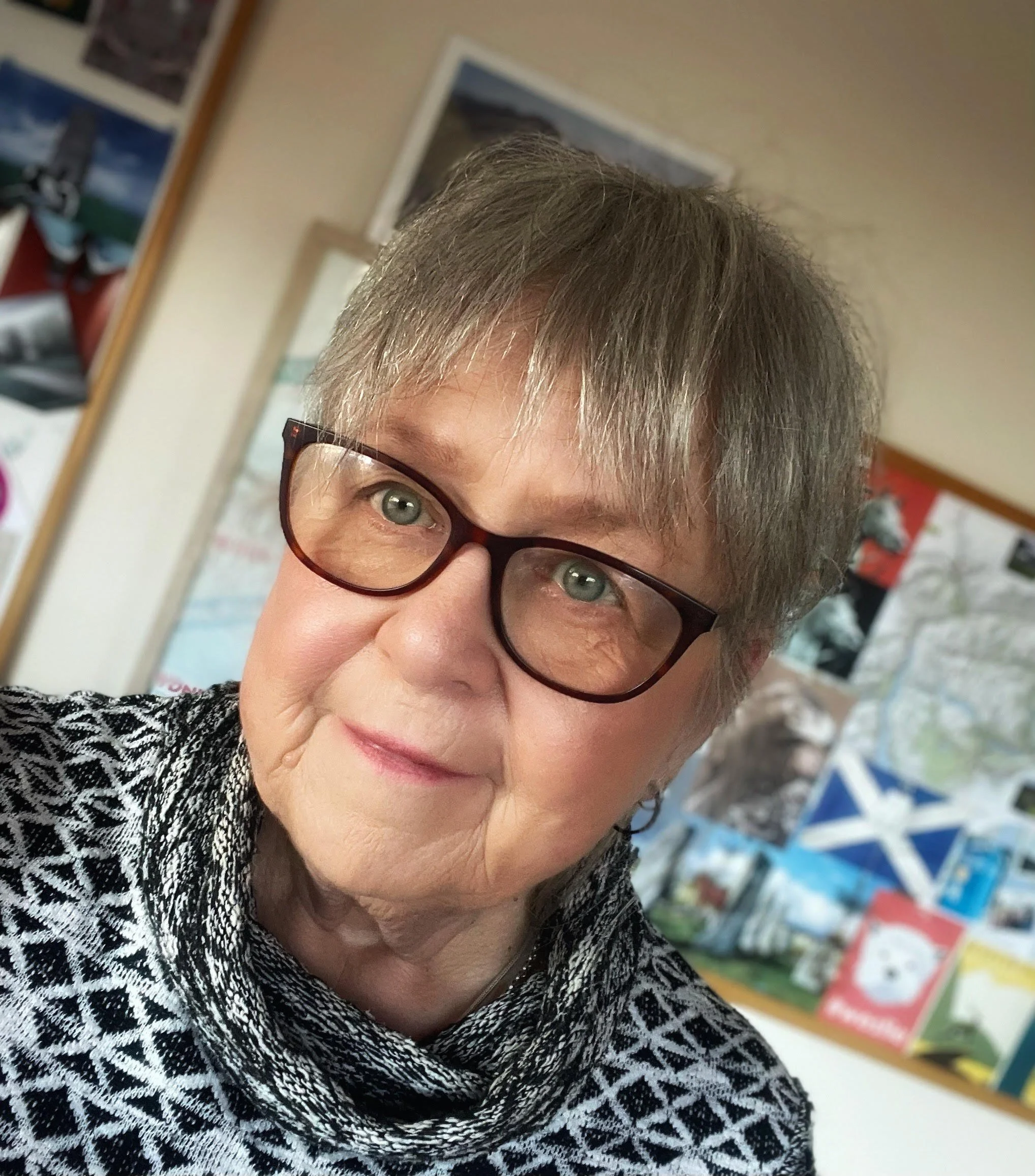 A portrait of a woman with short gray hair and glasses, wearing a black and white patterned scarf, standing in a room with a bulletin board in the background.