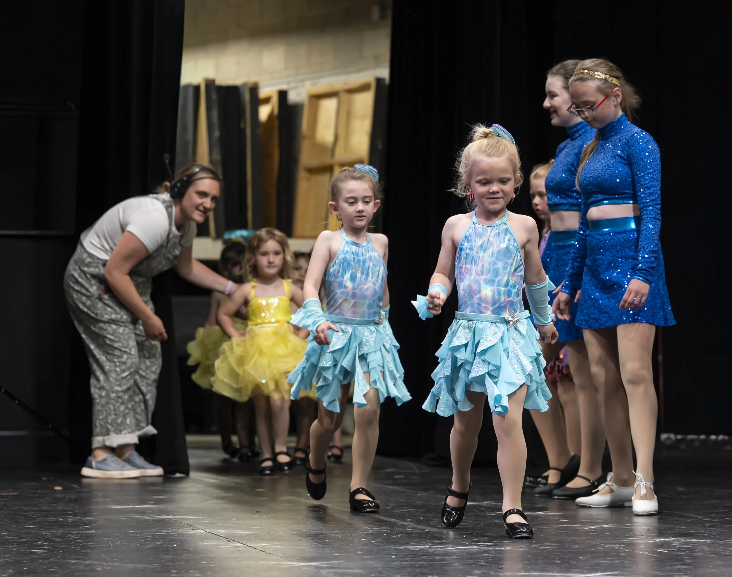 A woman guiding young girls onstage at a dance recital