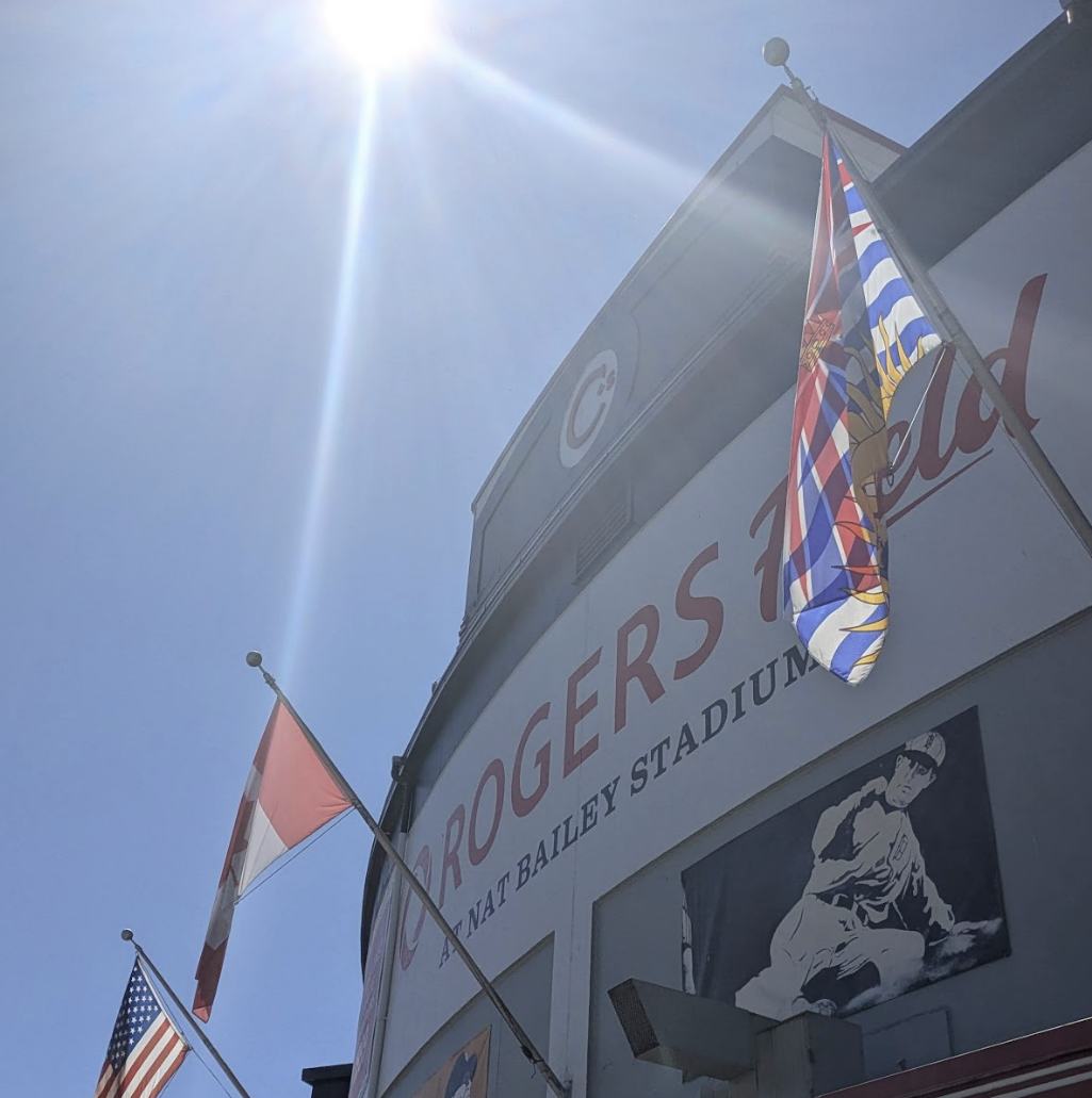 View of a stadium exterior with flags, including British Columbia Flag, Canadian flag, and American flag, against a bright sunlit sky, showing part of a sign that reads 'Canadians' at Nat Bailey Stadium. Designed by Sydenham Marketing.
