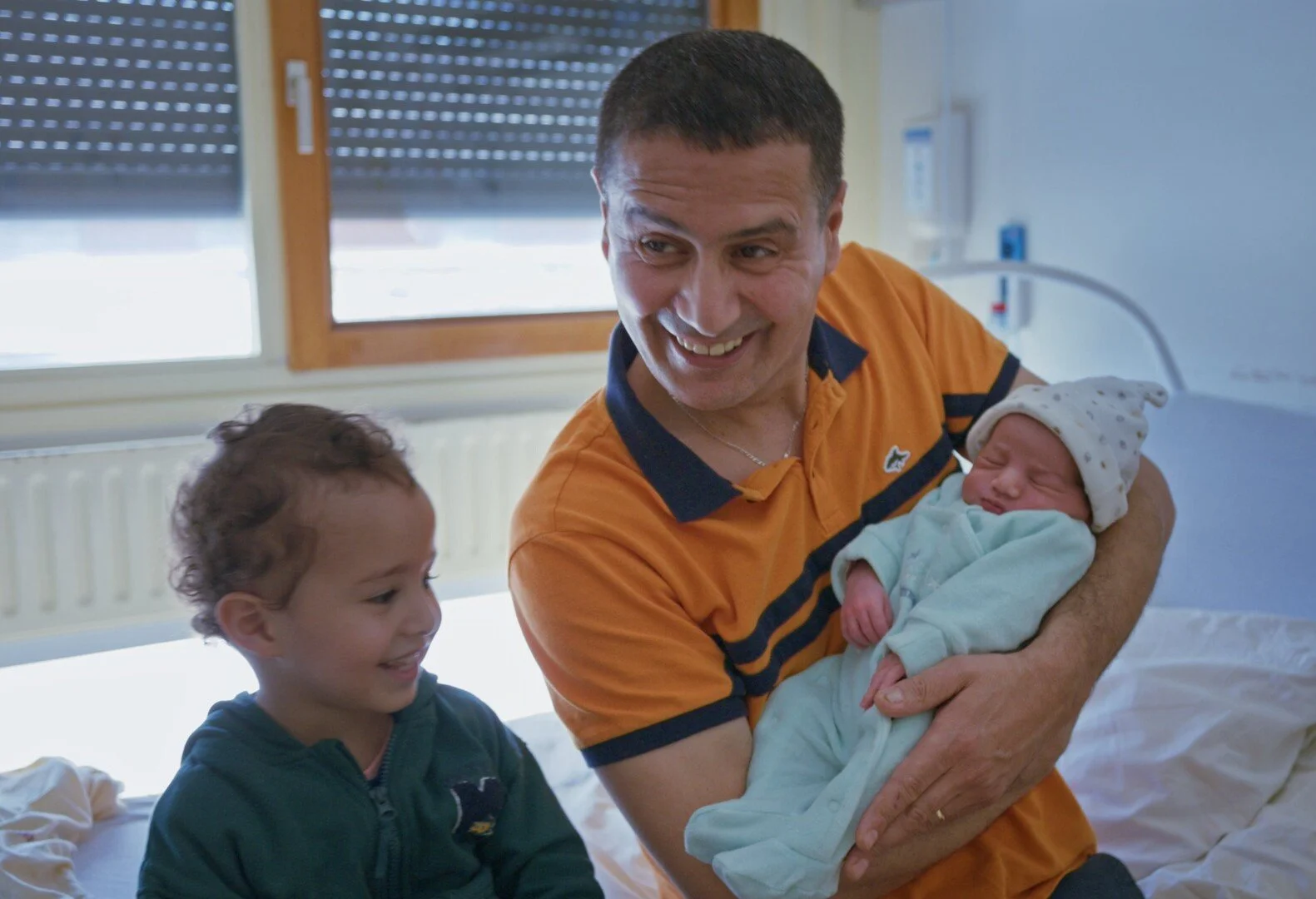 A man holds a newborn baby, showing the baby to a toddler sitting next to him