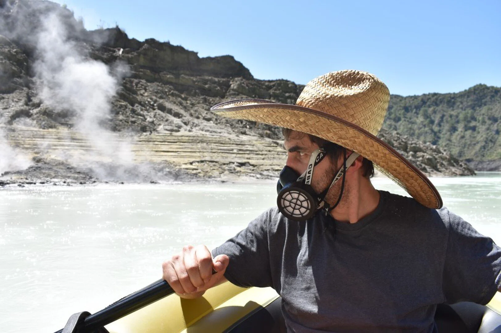 A man wears a sombrero and a gas mask, while rowing a canoe in a volcanic landscape