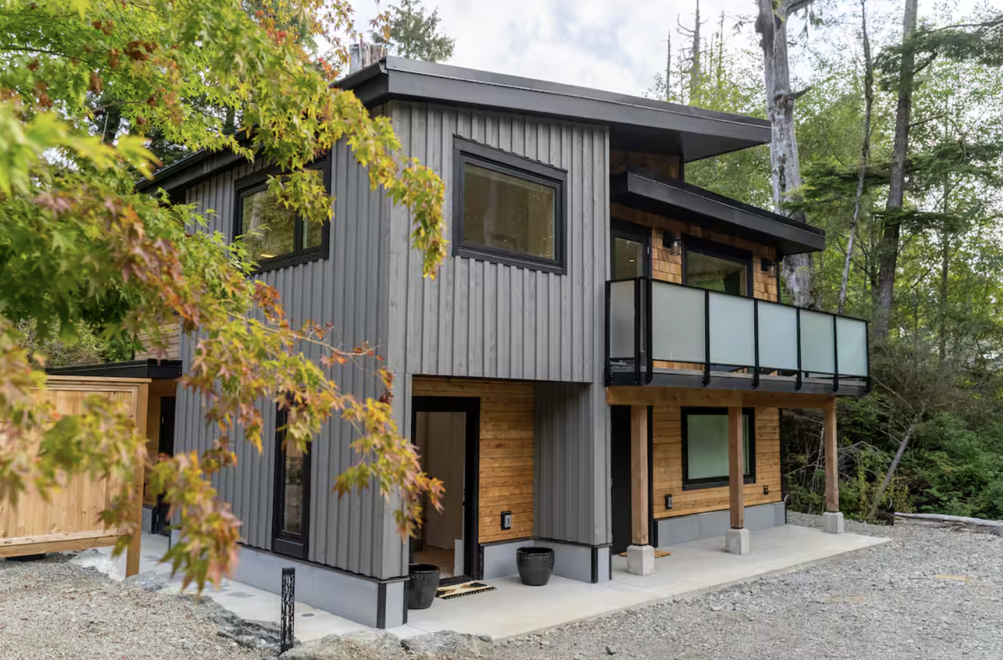 Modern two-story house with wood and metal siding, large windows, a balcony, surrounded by trees and greenery.