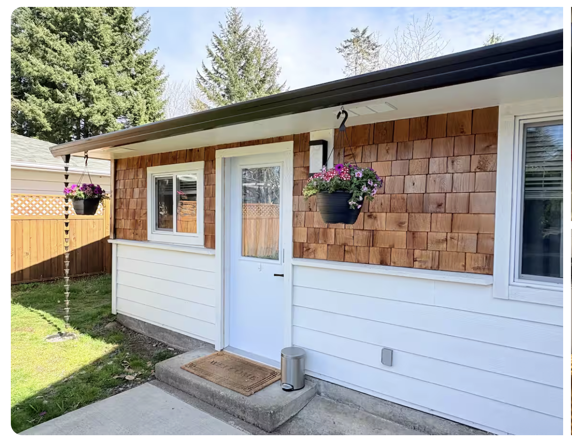 Front door of a house with hanging flower baskets, a doormat, and a small metallic trash can, with a wooden fence and trees in the background.