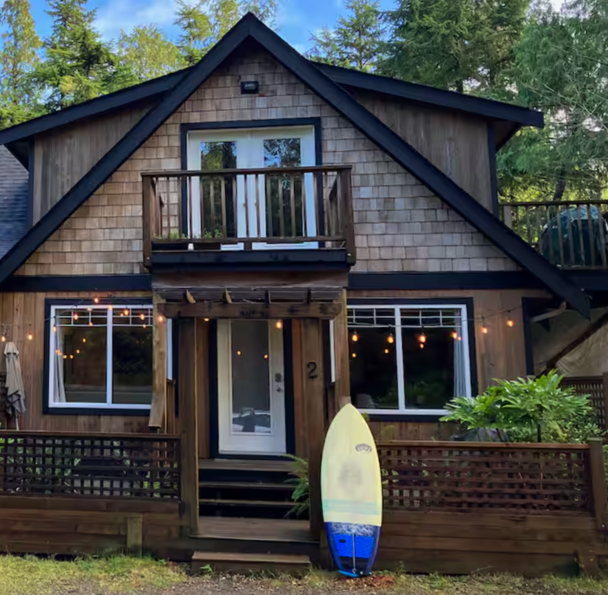 Two-story house with wooden siding, large front windows with string lights, a small balcony on the second floor, and a surfboard leaning against the front porch railing, surrounded by trees.