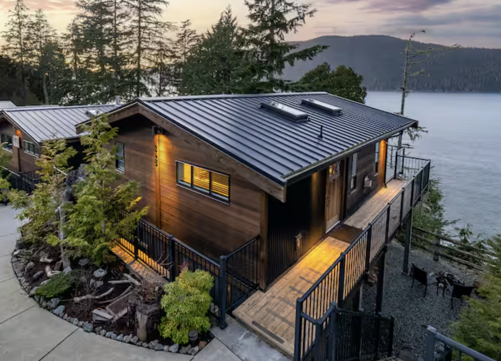 A modern house with a wooden exterior and a metal roof, overlooking a body of water and surrounded by trees during the evening.