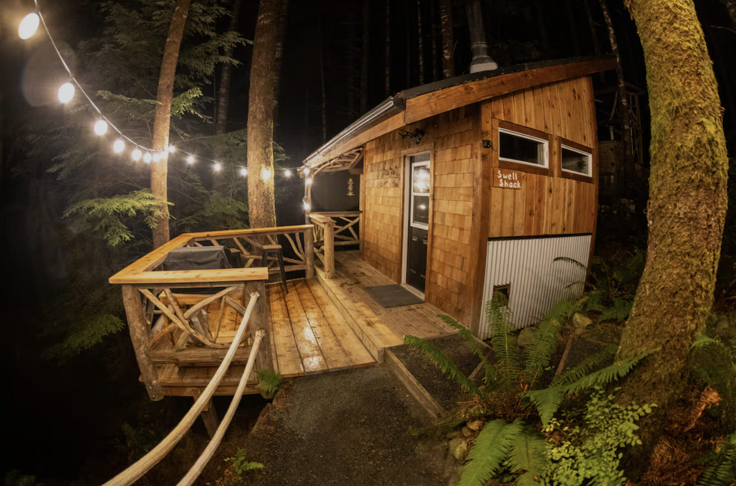 Wooden tiny house labeled 'Swell Shack' on a deck in a forest at night, lit by string lights, with trees and ferns surrounding it.
