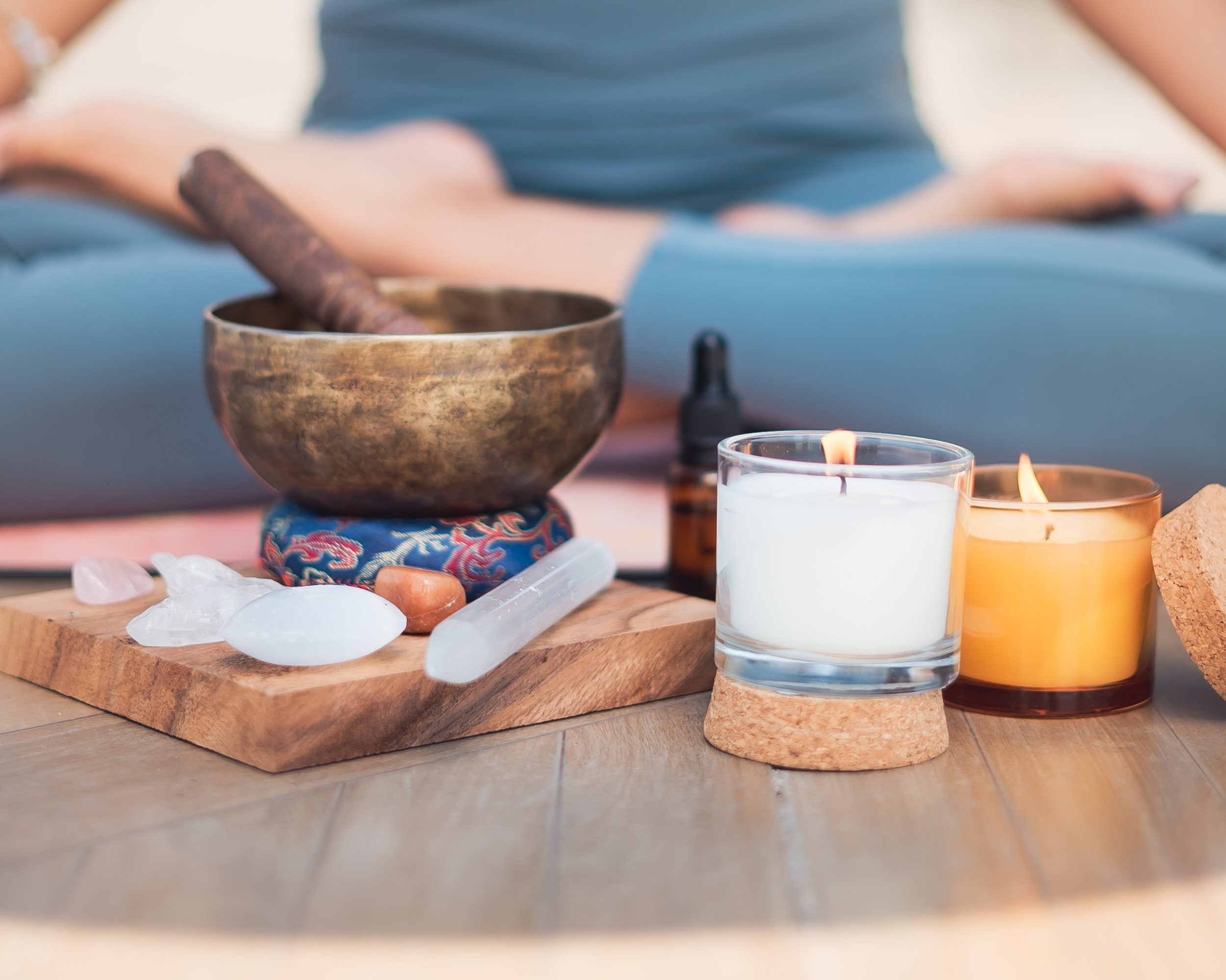 A crystal singing bowl with a wooden striker, surrounded by various crystals, candles, and essential oil bottles on a wooden surface, with a person seated in a cross-legged meditation pose in the background.
