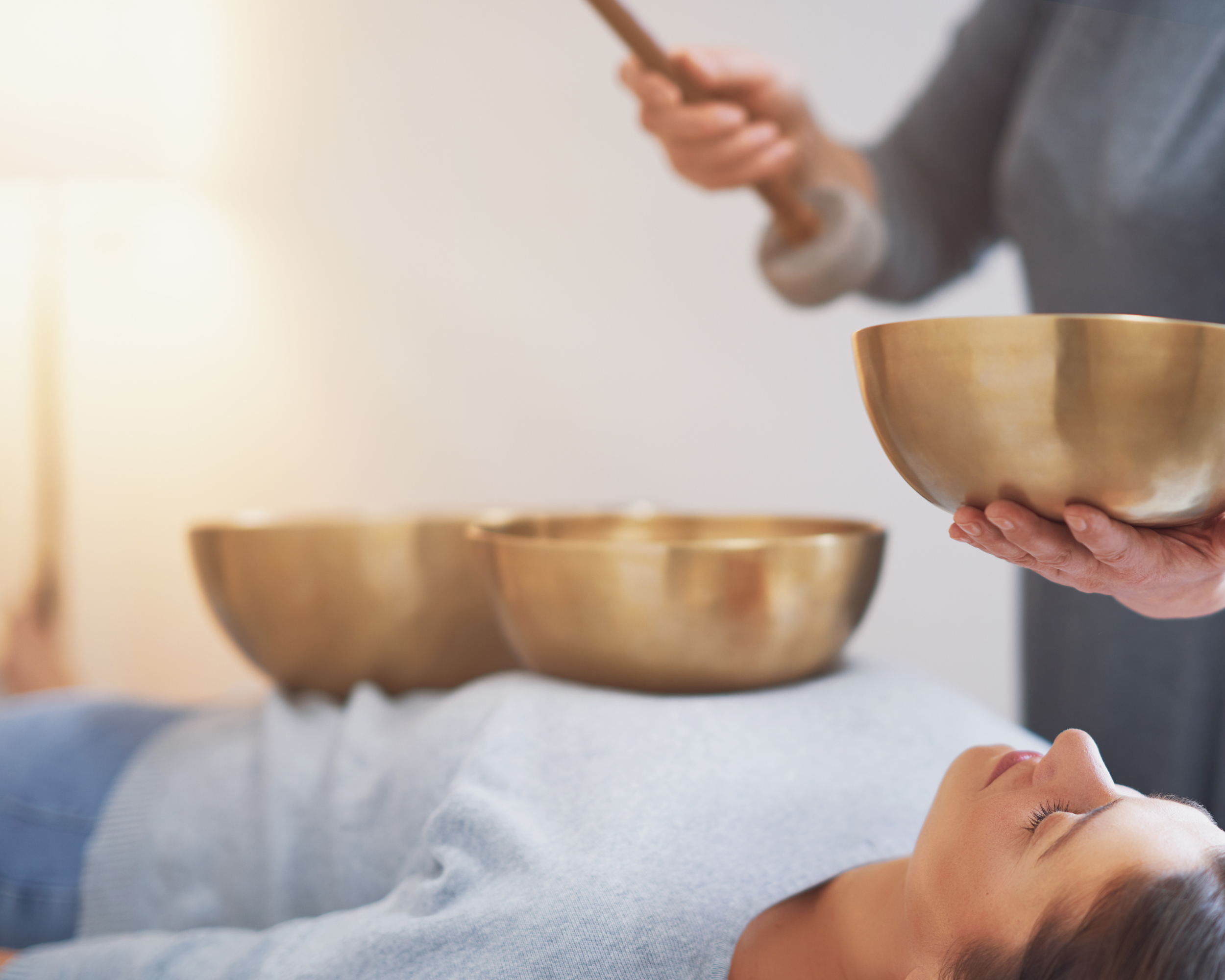 Person lying down with three singing bowls on their chest, while another person holds a fourth bowl and a mallet, preparing for sound therapy session.