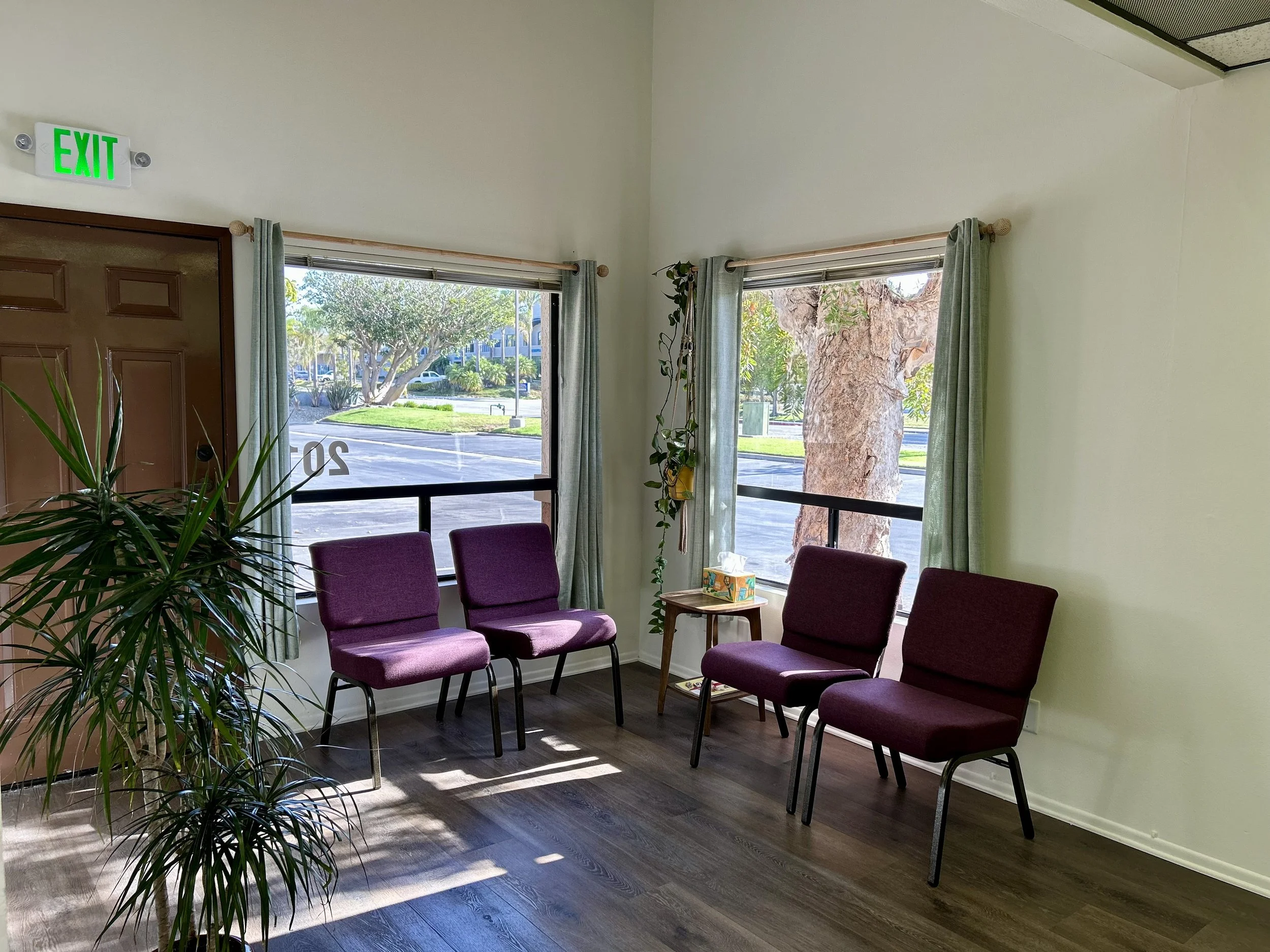 A waiting room with five purple chairs arranged in a semi circle. There is a table with a colorful tissue box and magazines. Green curtains frame large windows showing trees and parked cars outside. An exit sign is mounted above a wooden door.