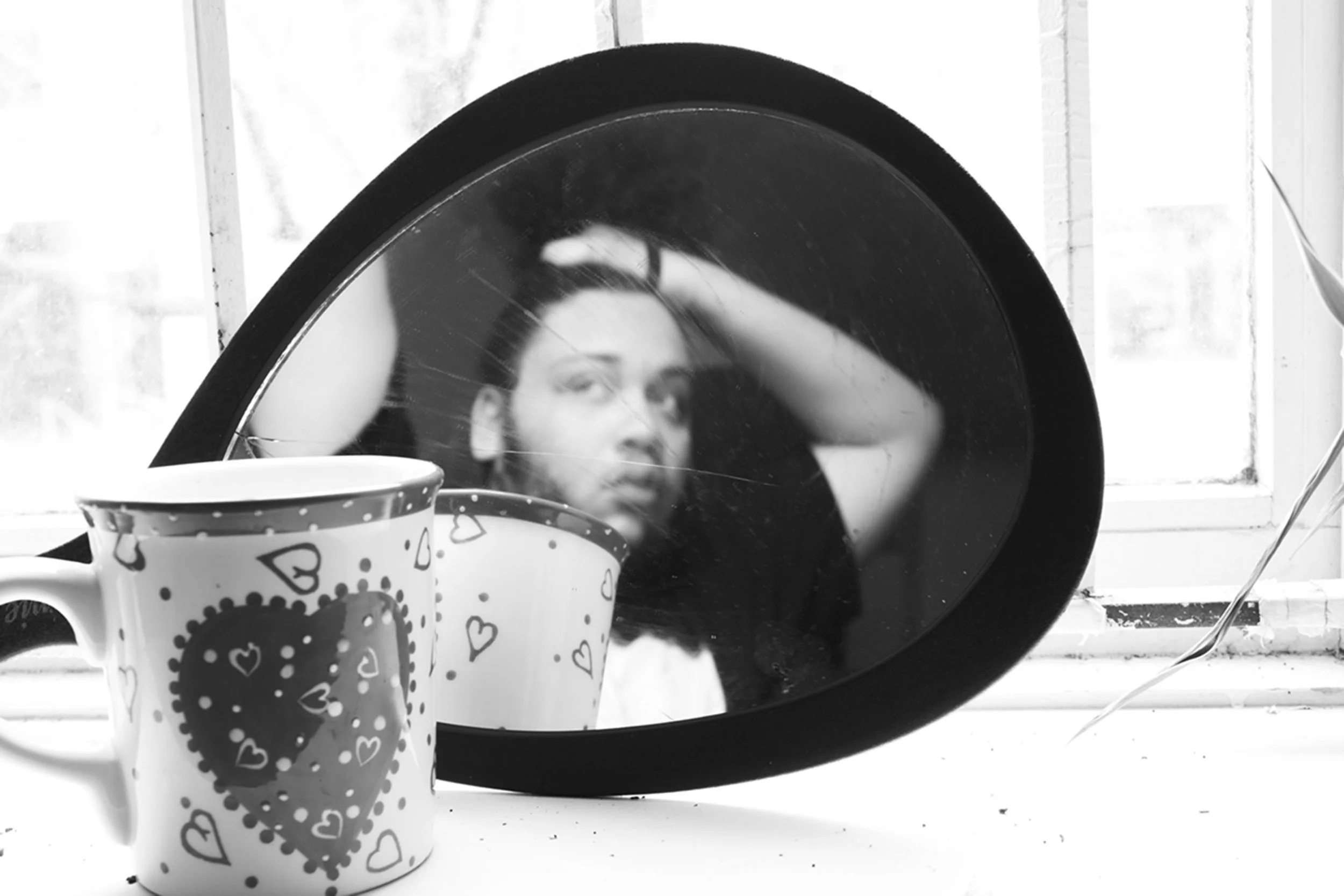 Black and white photo of a woman looking into a mirror, with three mugs decorated with hearts and polka dots in the foreground. The woman is touching her head with her right hand and has dark hair.