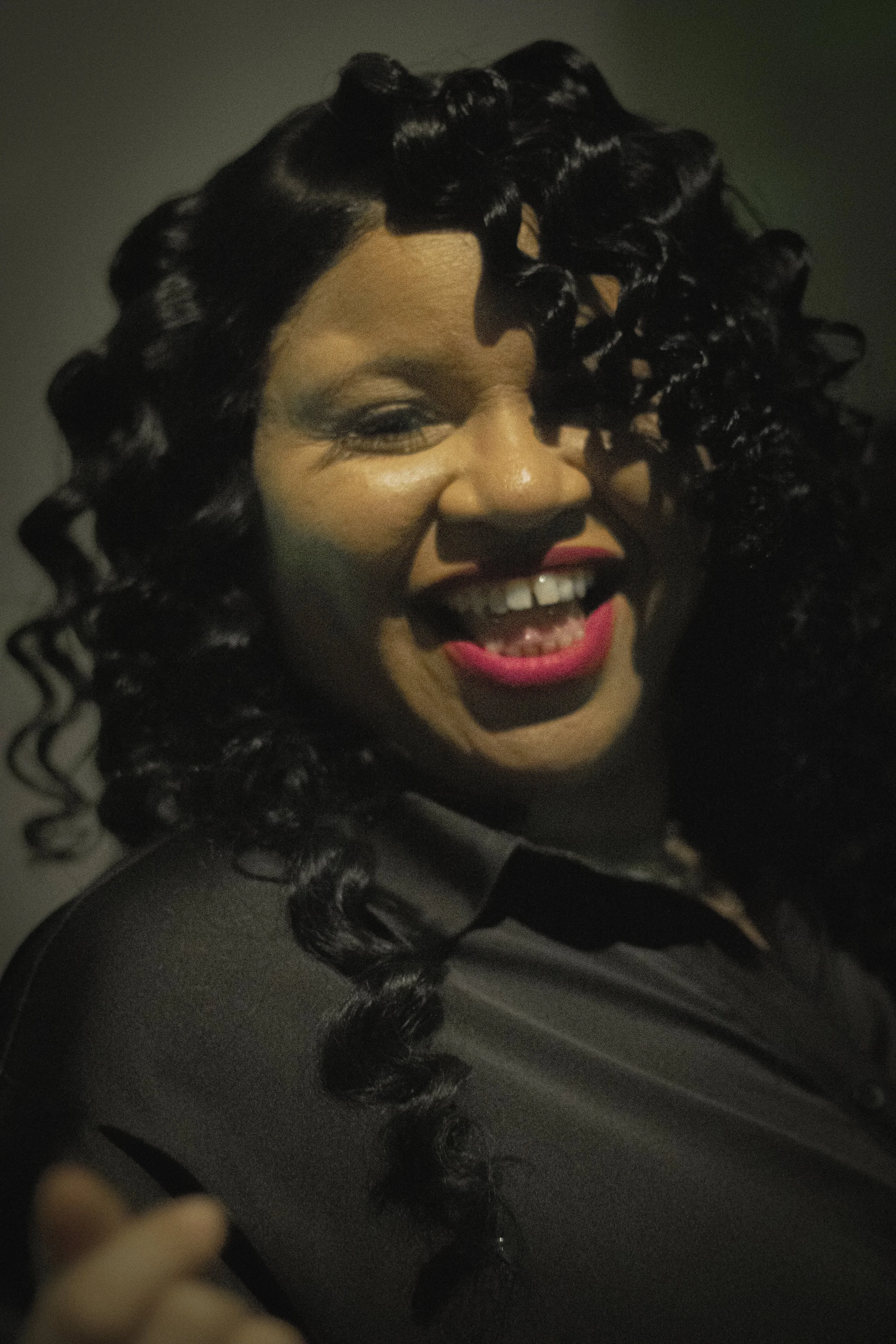 A woman with dark dark curly hair and makeup smiling with lipstick, partially visible in a dark setting.