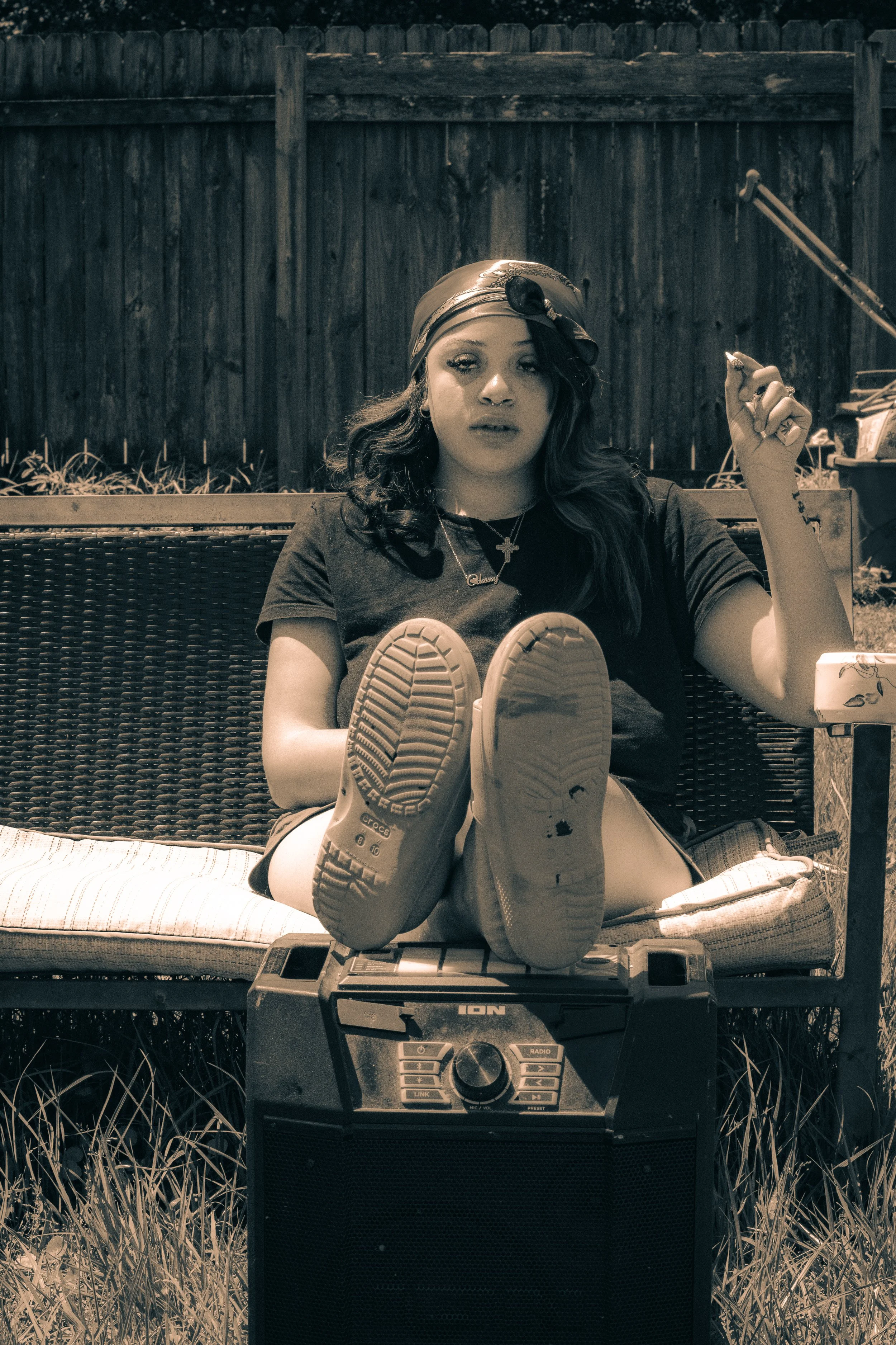 Black and white photo of a young woman sitting with her legs stretched out in front of her on a wicker outdoor sofa. She is wearing a headscarf, a necklace with a cross, and a black t-shirt. She is holding a cigarette in her right hand and has her left arm resting on her lap. The young woman is looking directly at the camera. In front of her is a large audio system or speaker on the ground, and to the right, a small table with miscellaneous items. The background features a wooden fence and some outdoor plants.