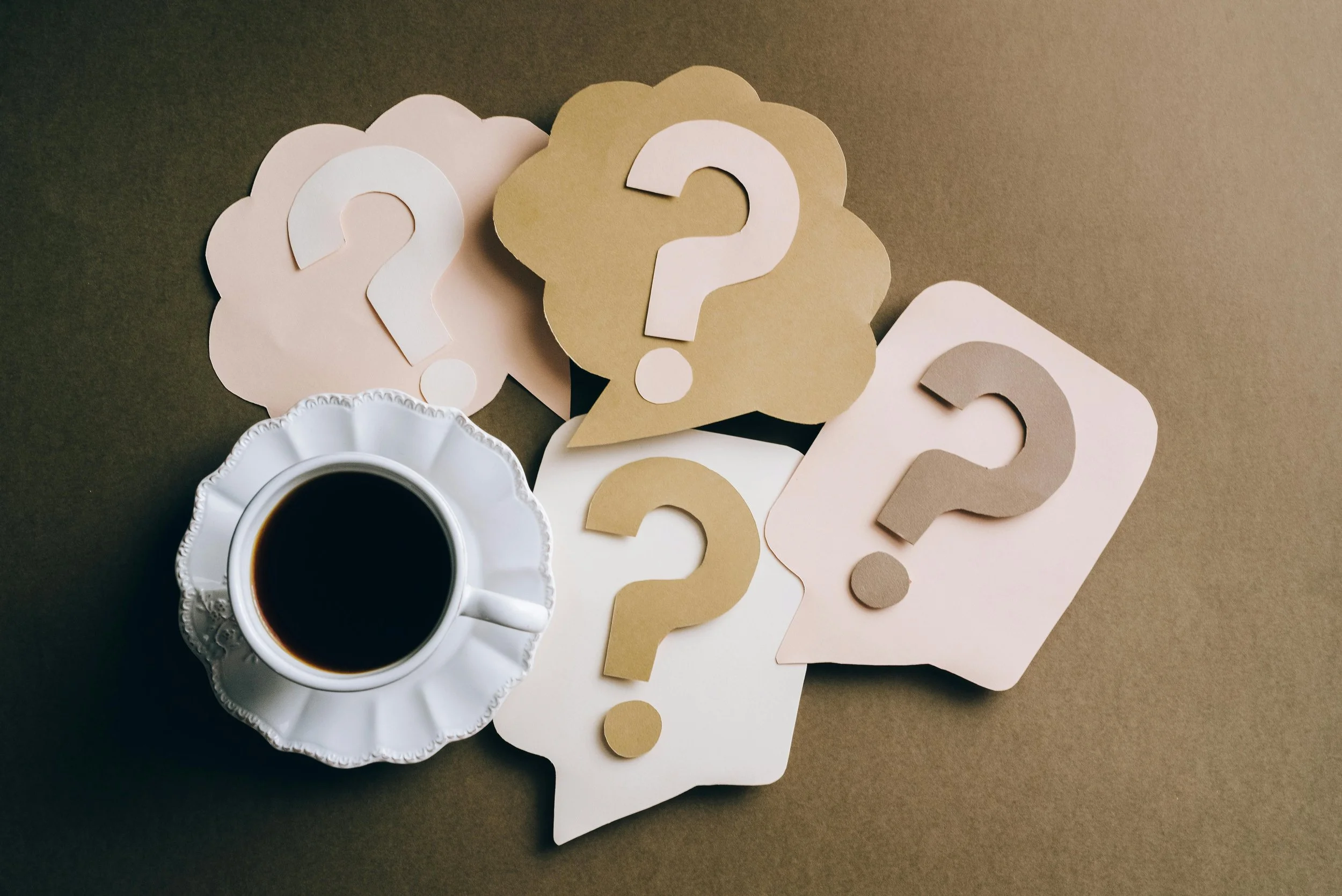 A cup of black coffee on a white saucer surrounded by paper speech bubbles with question marks in beige, white, and pink colors on a brown surface.
