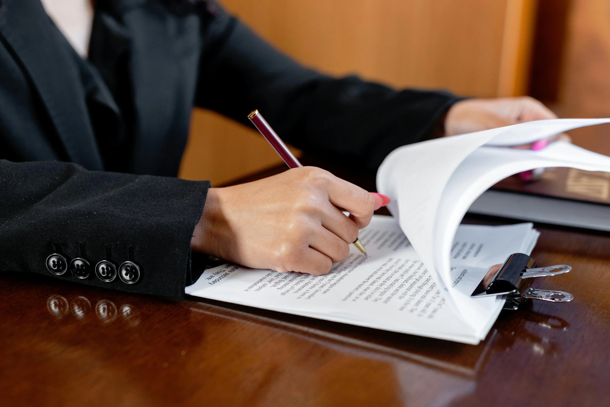Person in black suit reviewing and signing documents on a wooden table with a binder clip holding papers together.