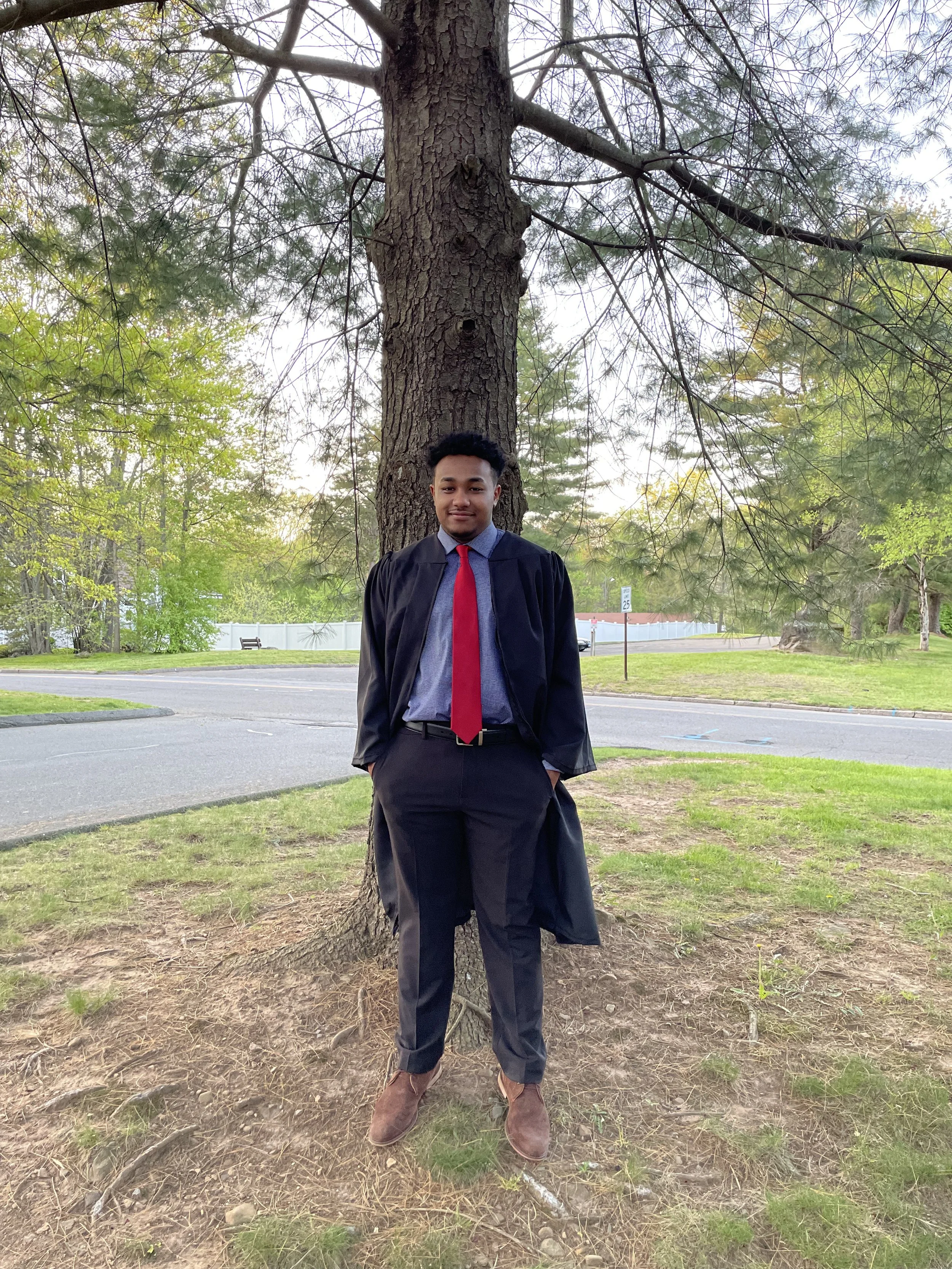 Young man in graduation gown and cap, standing outdoors near a large tree, smiling, with a street and grassy area in the background.