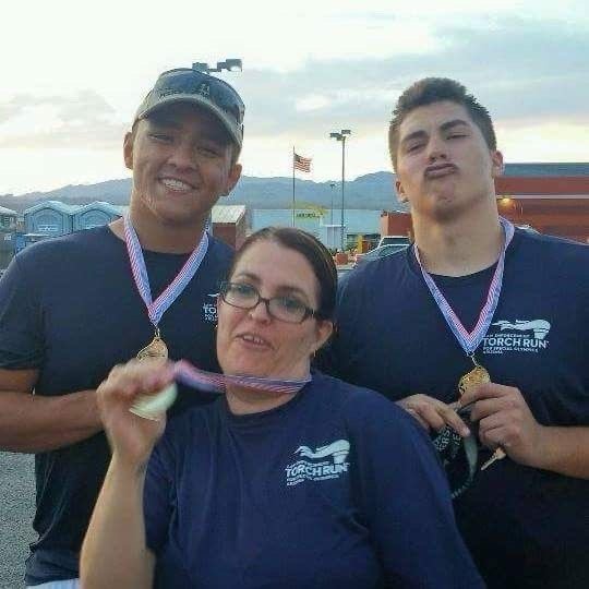 Three people wearing medals and matching navy T-shirts, standing outdoors with a cloudy sky and buildings in the background, celebrating at an event.