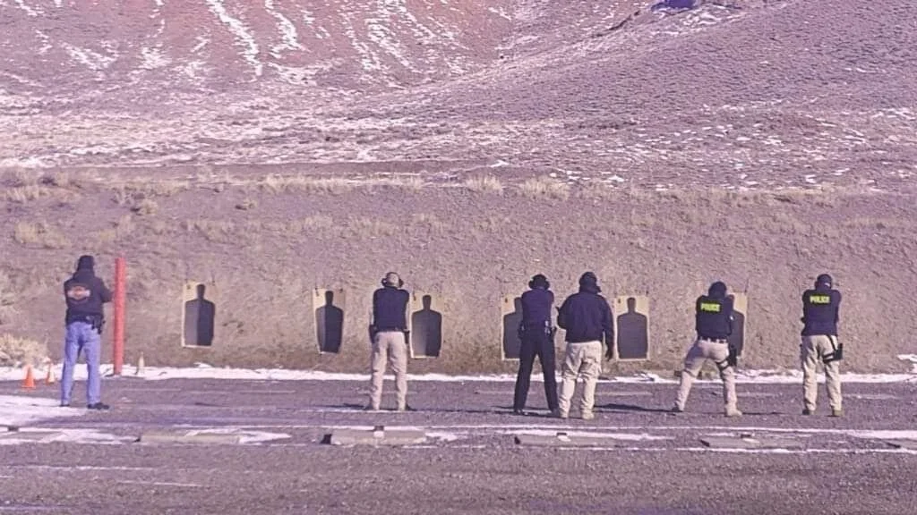 Seven individuals, some in police vests, stand at a shooting range, aiming at multiple silhouette targets on a hillside.