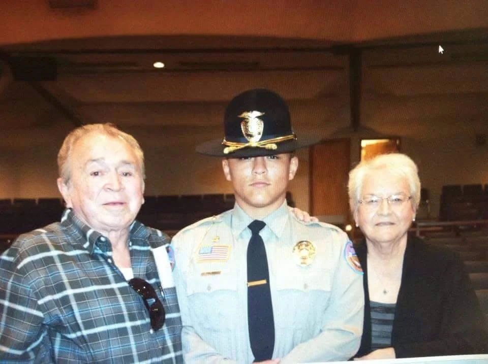 Three individuals standing together indoors: a senior woman with white hair and glasses on the right, a police officer in uniform in the center, and an older woman with gray hair and glasses on the left wearing a plaid shirt.