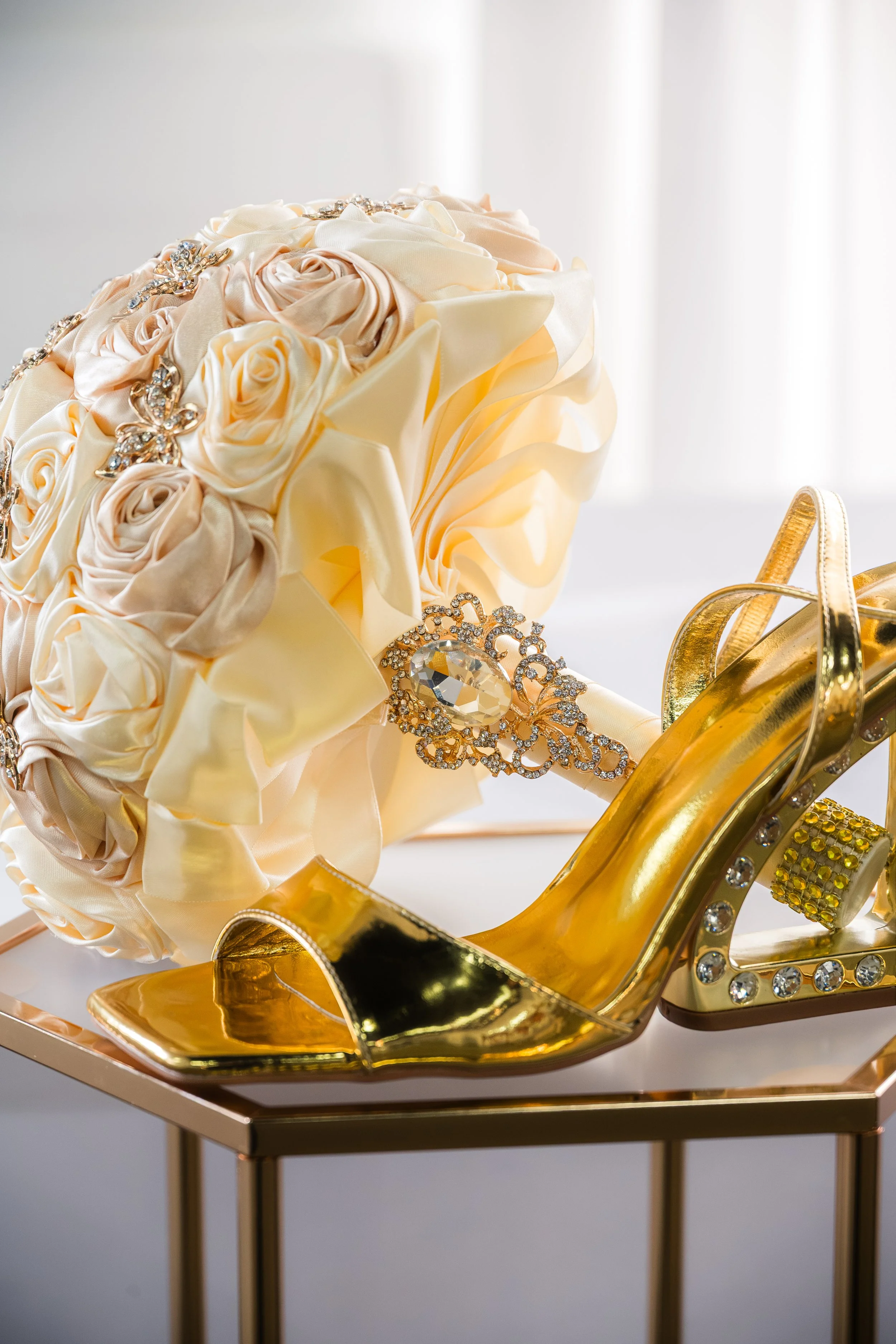 Golden high-heeled shoe with rhinestone embellishments displayed on a stand, next to a bouquet of cream-colored satin roses with rhinestone jewelry resting on it.