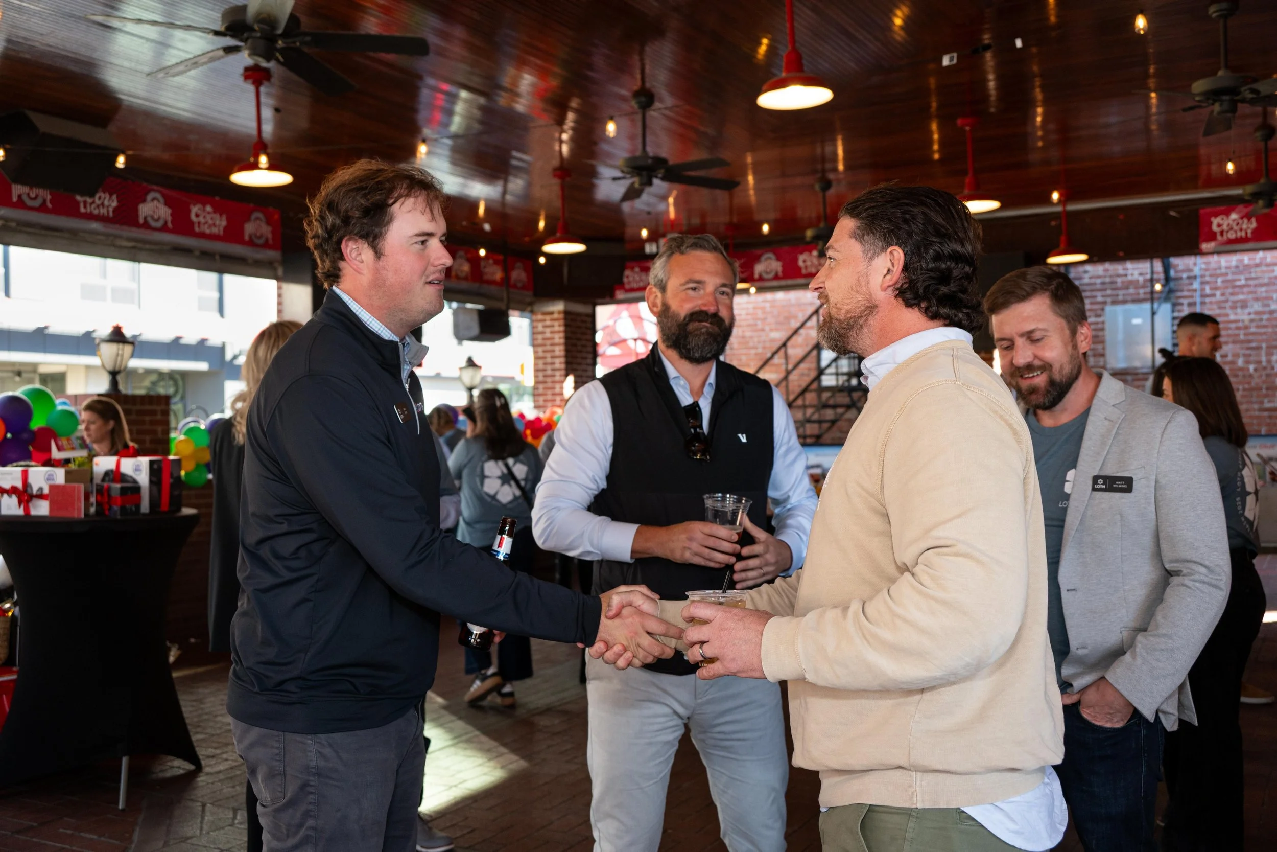 Four men in conversation at a social gathering or networking event inside a venue with brick walls, wooden ceiling, and red accents, including hanging lights and umbrellas.