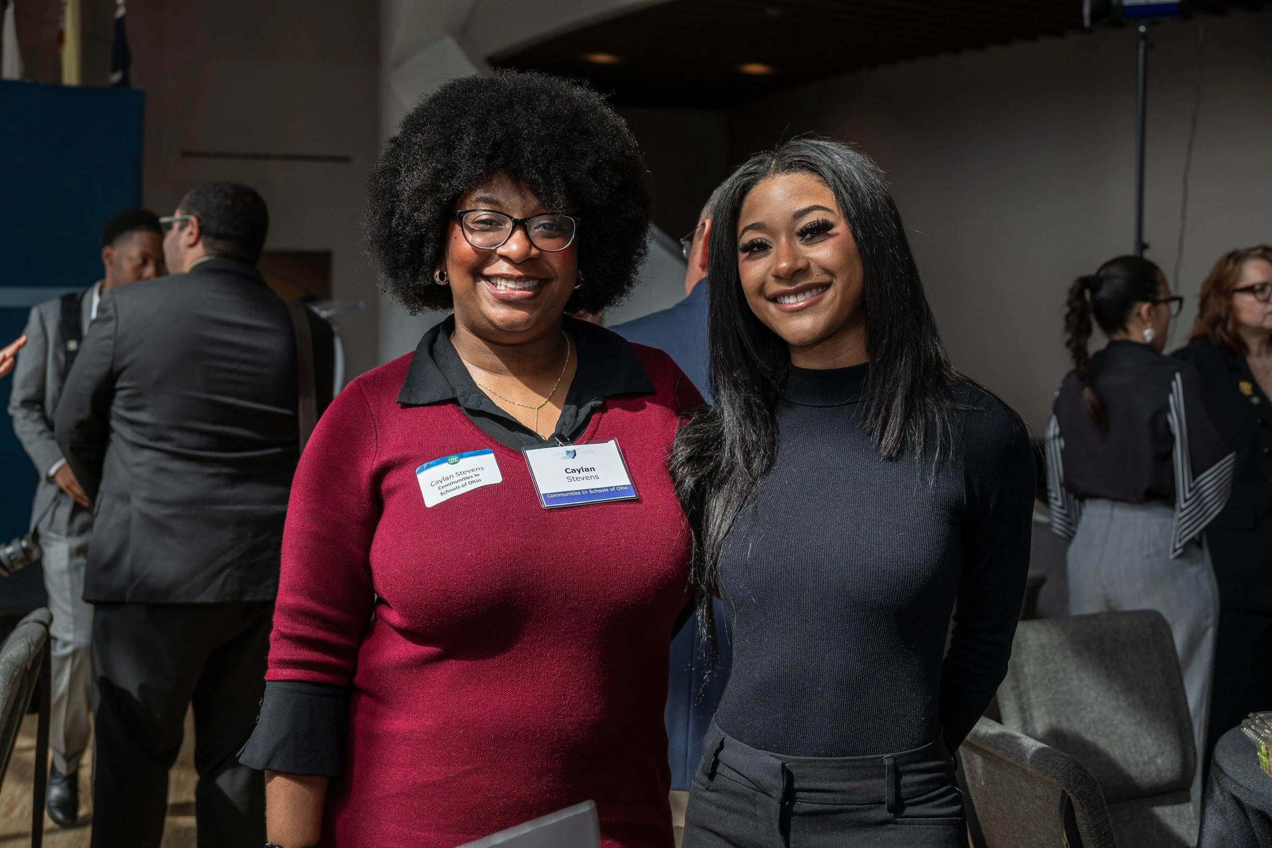 Two women smiling at an indoor event, wearing name tags, surrounded by other attendees.