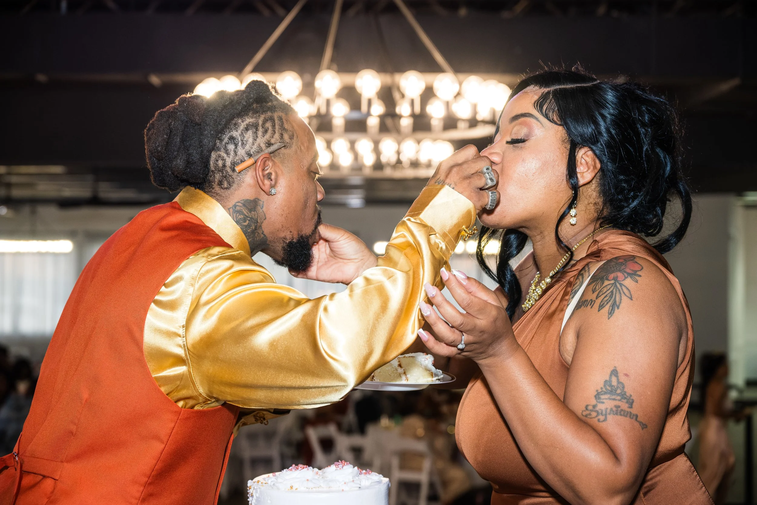 A man with dreadlocks and tattoos on his neck and hand is feeding a woman with black hair and tattoos on her arm a piece of cake at a celebration or party.