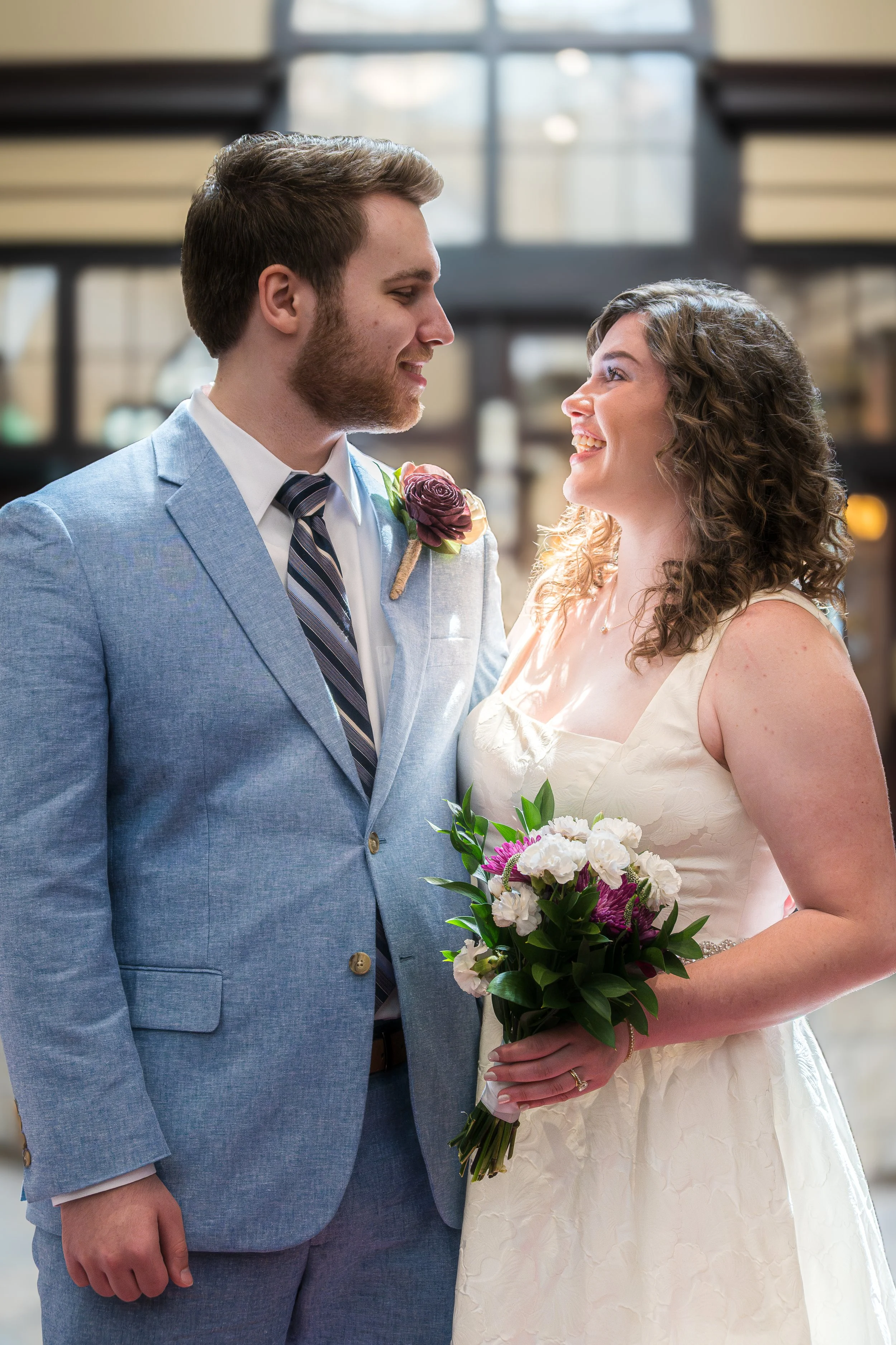 A smiling couple is standing close together, facing each other indoors. The man is wearing a light blue suit with a white shirt and a striped tie, with a boutonniere on his lapel. The woman is wearing a white dress, holding a bouquet of white, pink, 