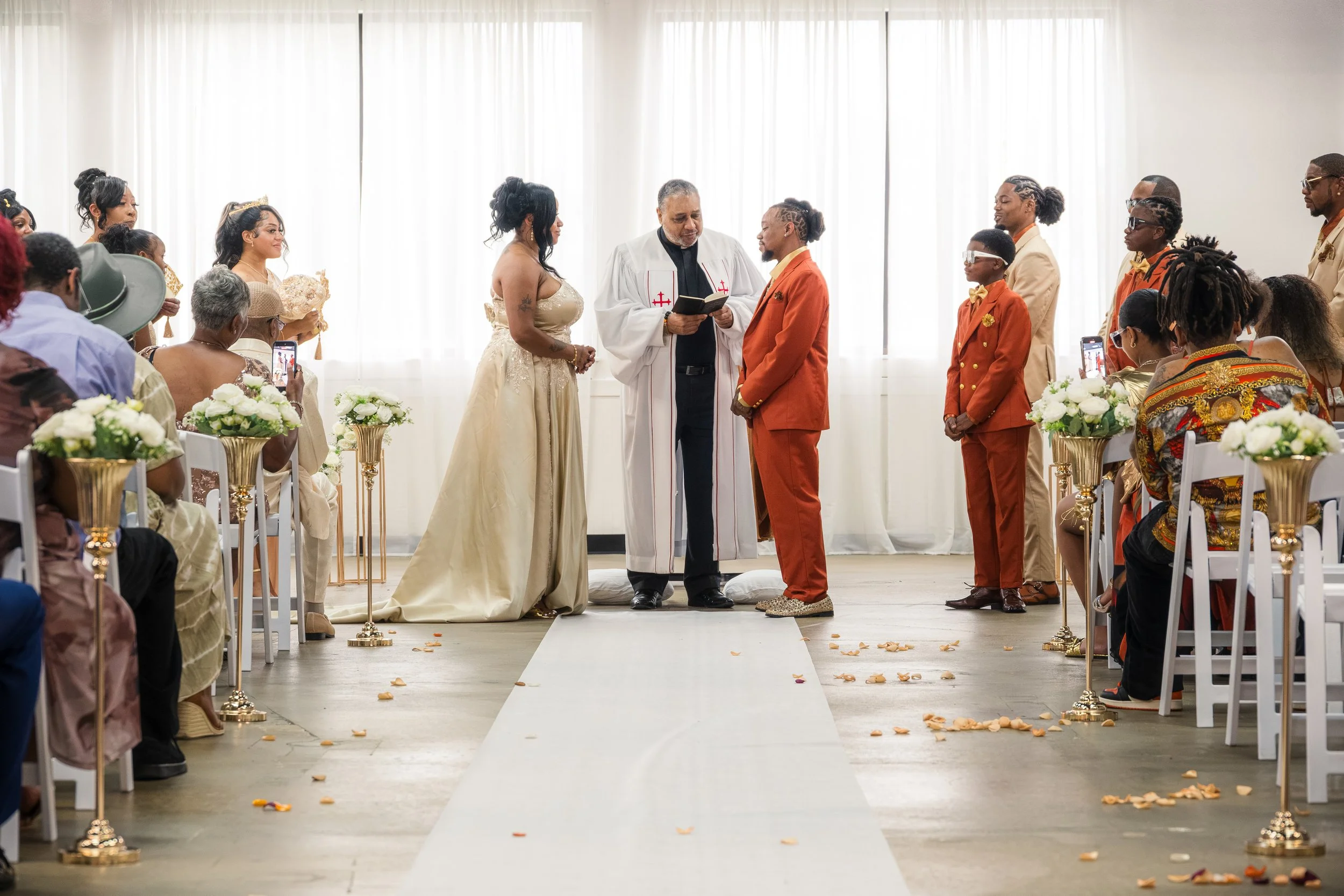 A wedding ceremony with a diverse group of people, including the bride and groom, officiant, and guests, stationed in a bright room with white curtains. Guests are seated on either side of a white aisle decorated with scattered flower petals, and som