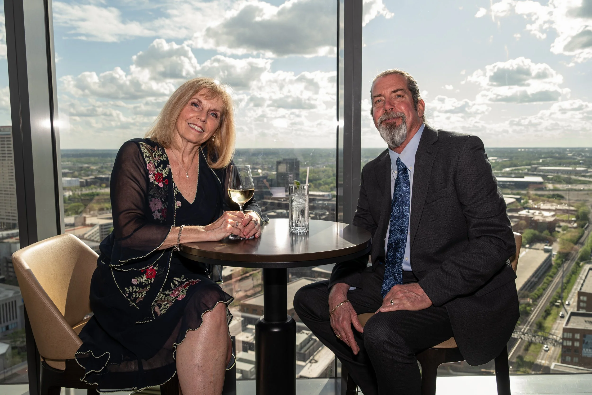 An older woman and man sitting at a round table in a high-rise building with a city view, smiling. The woman holds a glass of white wine, and the man has a glass of water. The woman wears a black dress with floral embroidery, and the man wears a gray