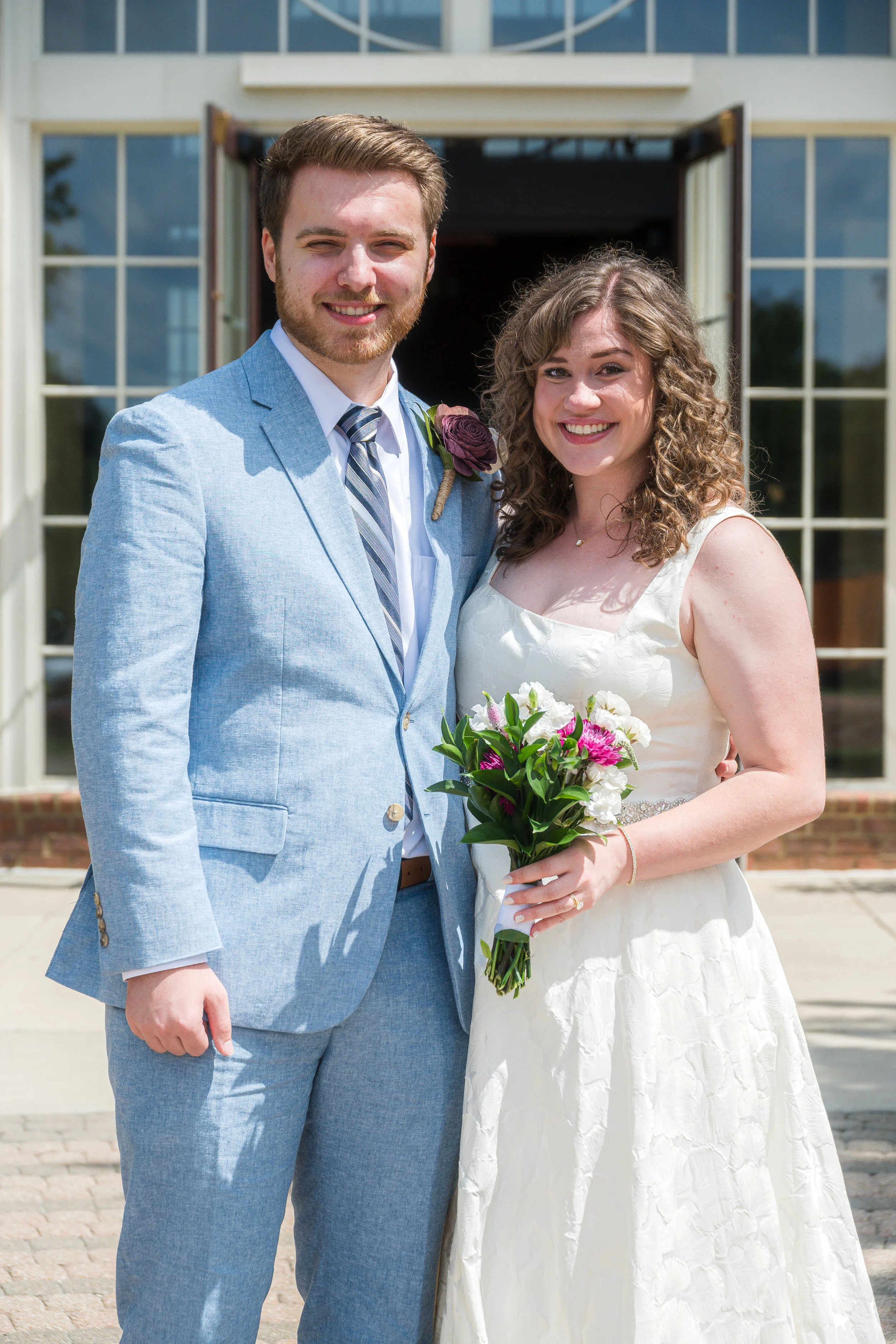 A smiling couple dressed in wedding attire standing outside in front of a building with large glass windows and open doors. The man wears a light blue suit with a striped tie and a brooch, while the woman wears a white wedding dress and holds a bouqu
