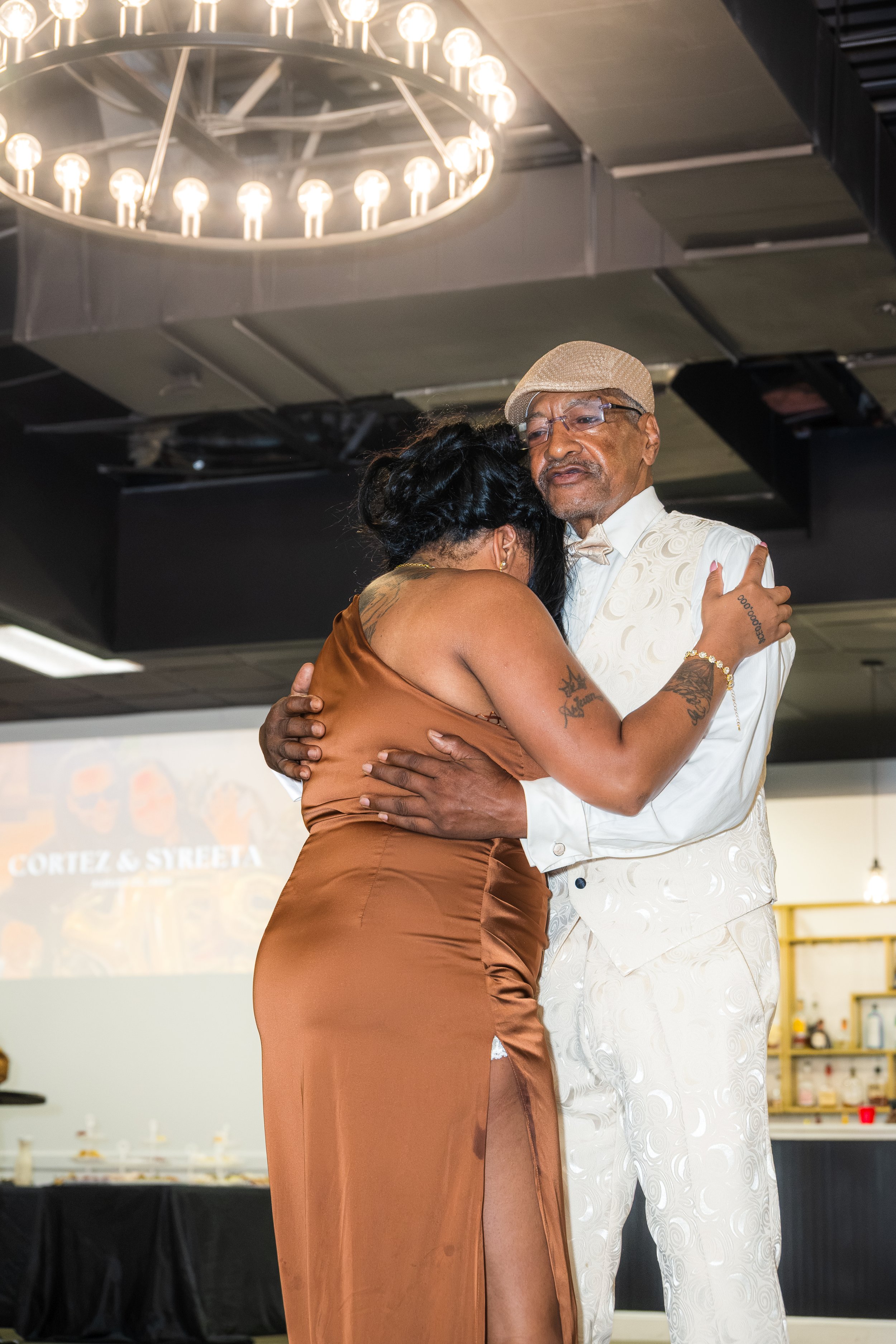 An elderly man and a woman are dancing closely together in an indoor event. The man is wearing a cream-colored vest and pants with a pattern, a beige flat cap, and glasses. The woman has tattoos on her arms and shoulder, and is wearing a bronze-color