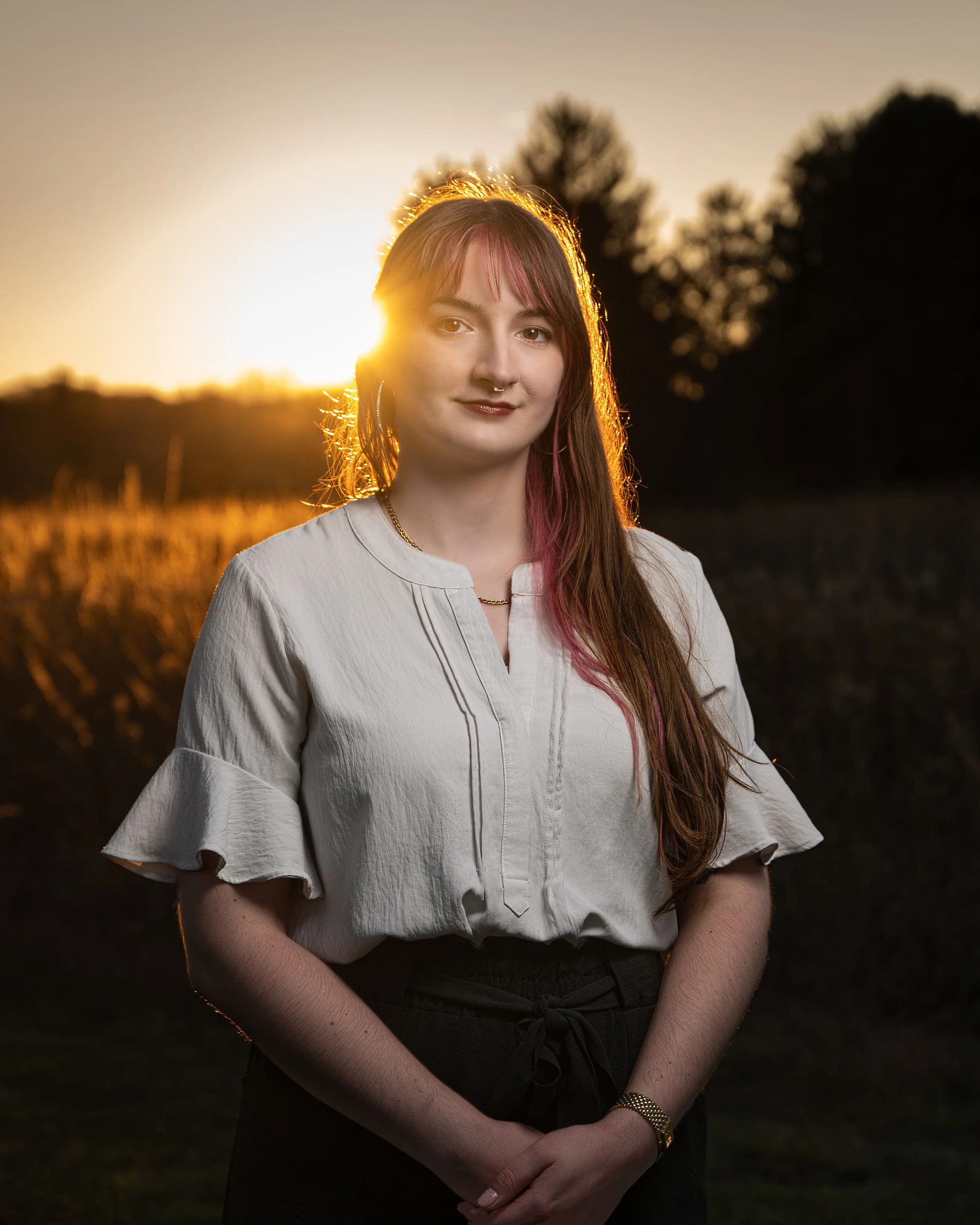 A young woman with long reddish-brown hair and pink streaks standing outdoors during sunset, wearing a white blouse with ruffled sleeves and dark pants, with a background of trees and a golden sky.