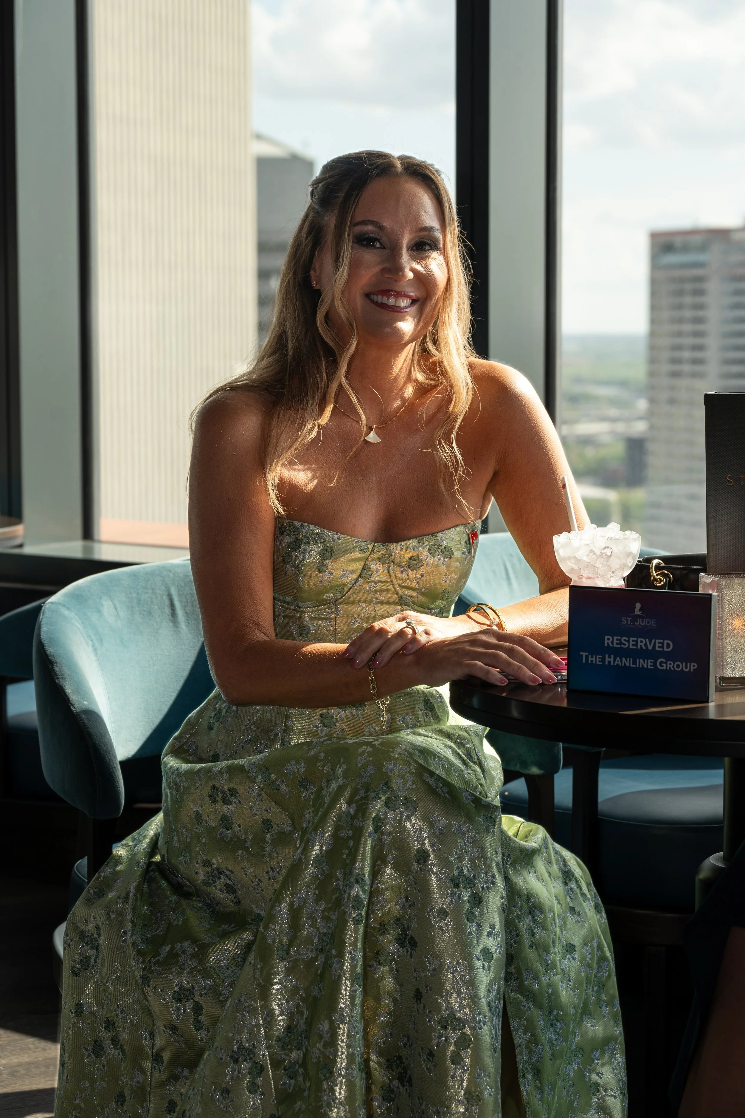 A smiling woman with wavy blond hair wearing a strapless, gold and green floral dress, sitting at a table with a drink and reserved sign, inside a modern building with large windows and cityscape view.