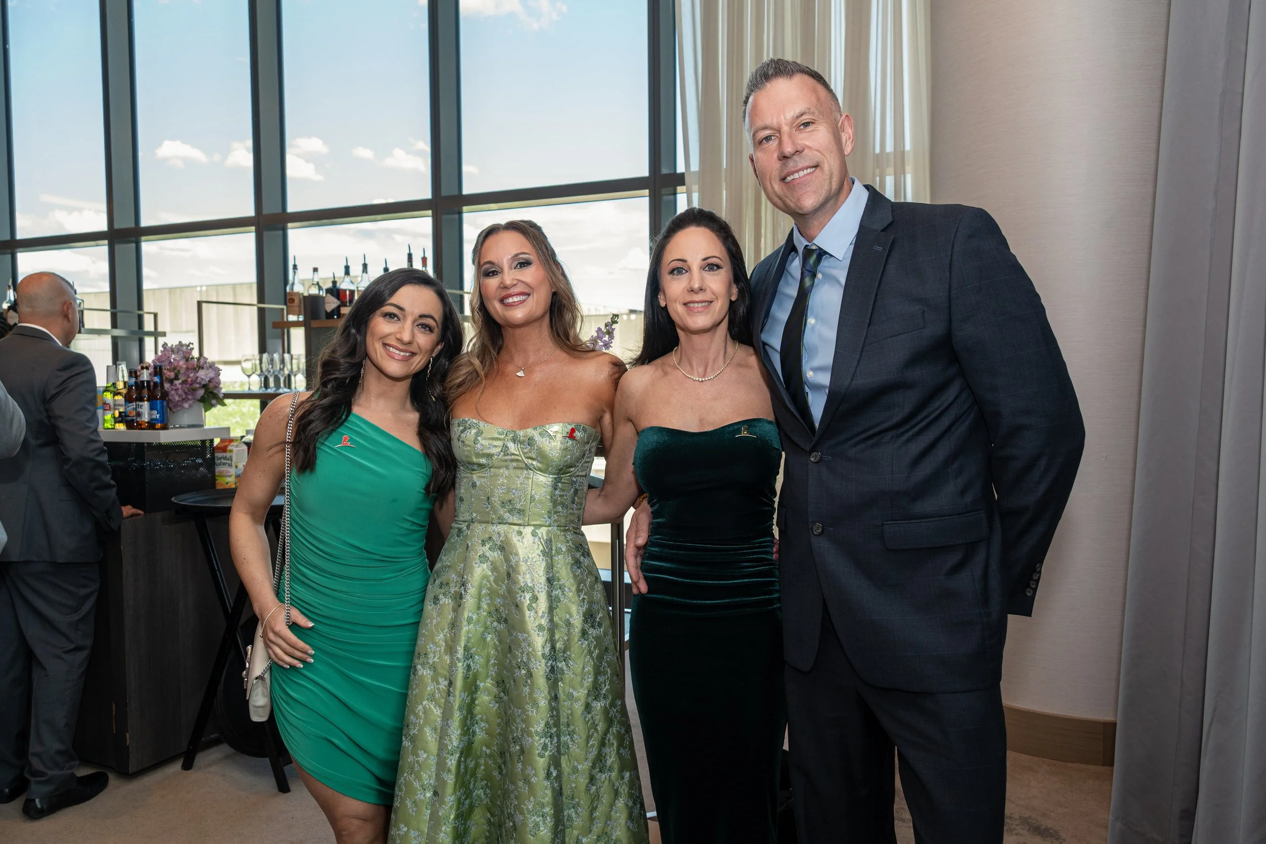 Four people, three women and one man, dressed in elegant attire, posing together at a social event in a bright room with large windows and a bar in the background.