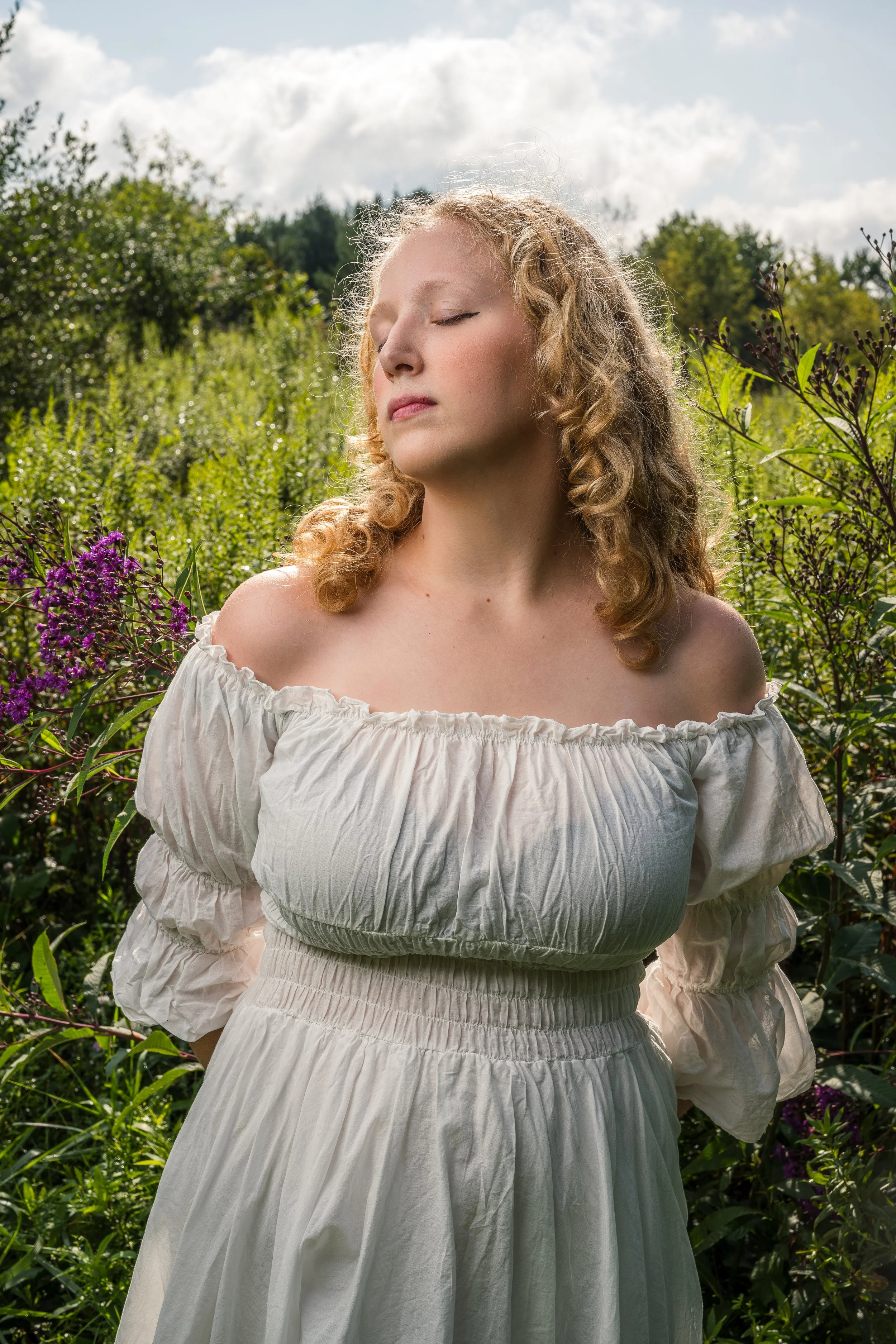 A young woman with curly blonde hair and closed eyes standing outdoors in a field with green plants and purple flowers, under a partly cloudy sky, wearing a white off-the-shoulder dress.