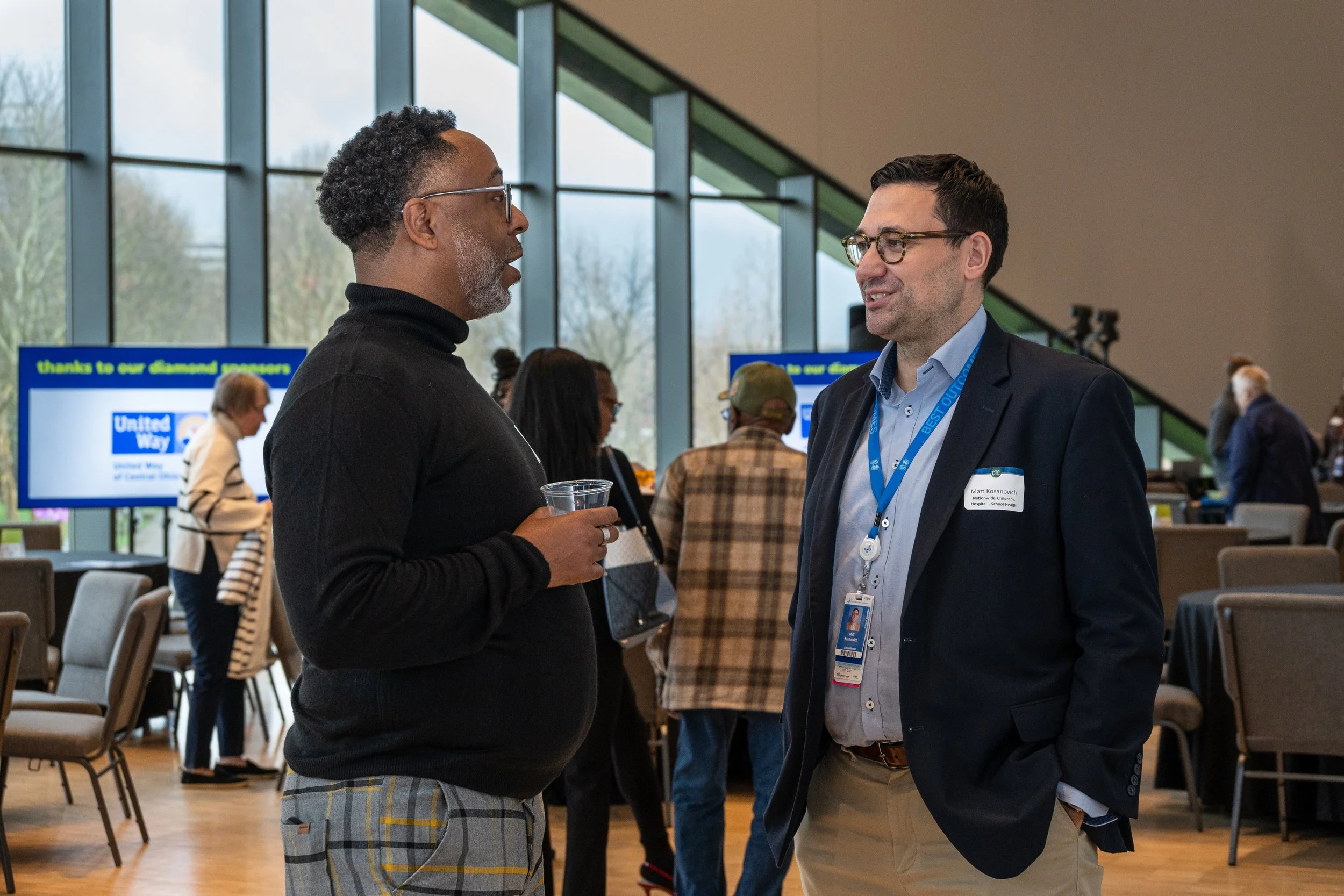 Two men are engaged in conversation at a professional event, one wearing a black turtleneck and glasses, and the other in a blazer with a name tag, in a spacious venue with large windows and people in the background.