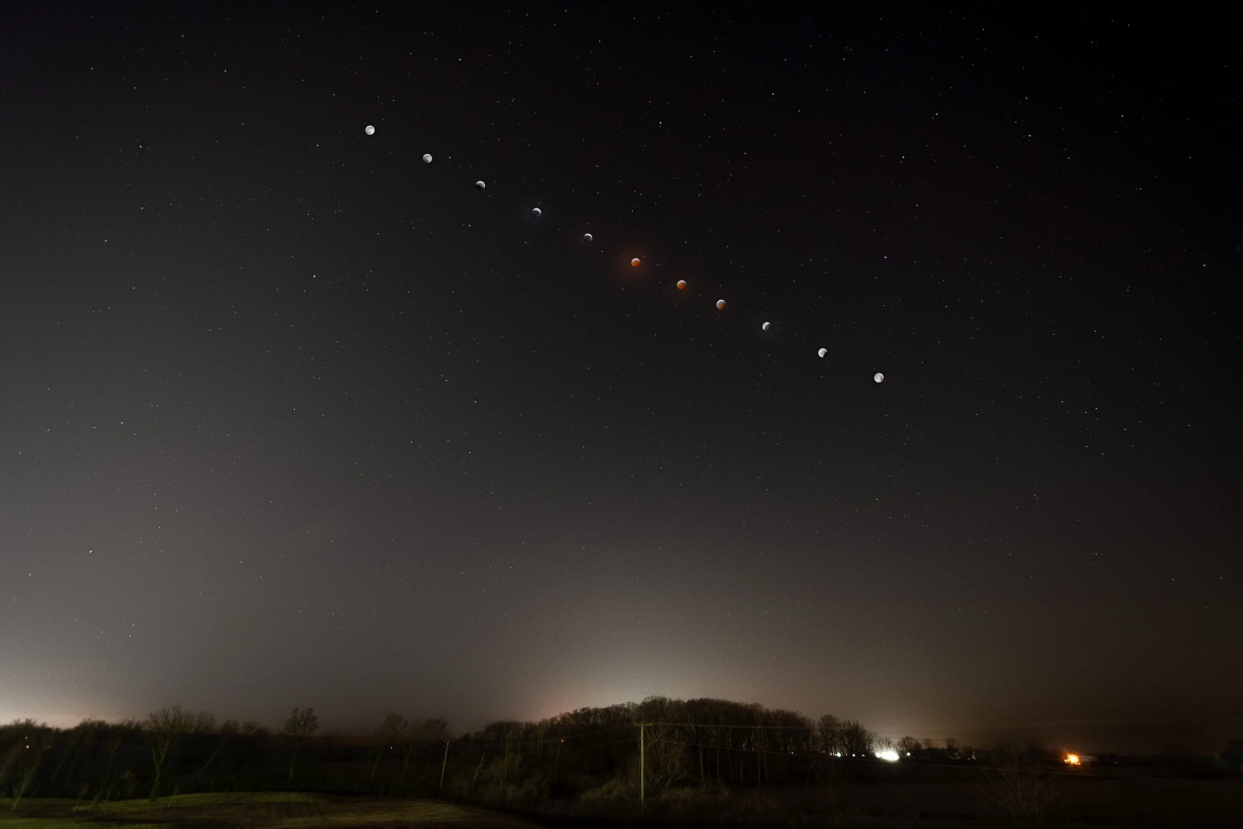 Night sky showing the moon in different phases from new moon to full moon aligned diagonally, with stars scattered across the sky and a landscape with trees and power lines at the bottom.