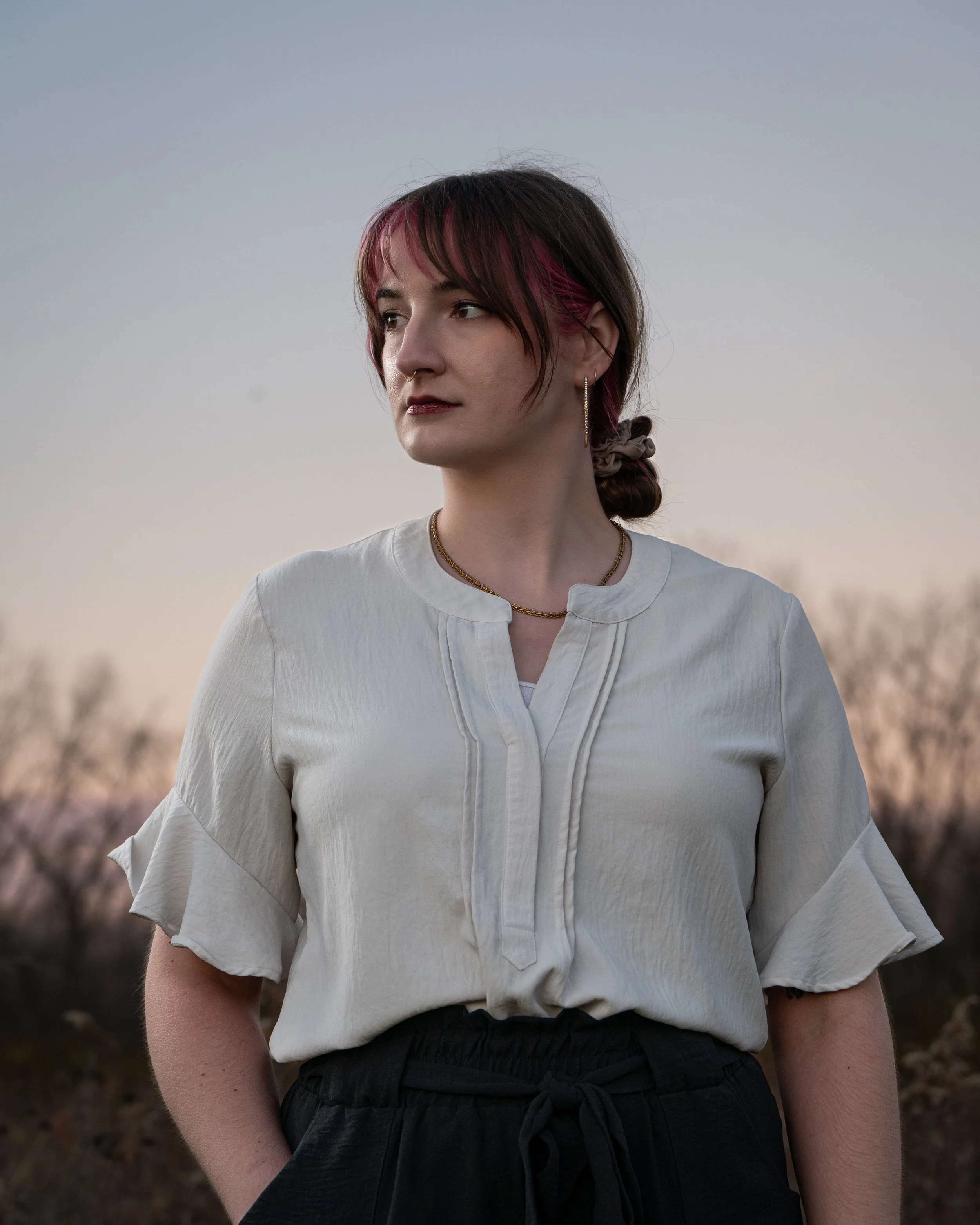 A woman with short hair that has pink highlights, standing outdoors during twilight, wearing a cream-colored blouse with ruffled sleeves and black drawstring shorts.