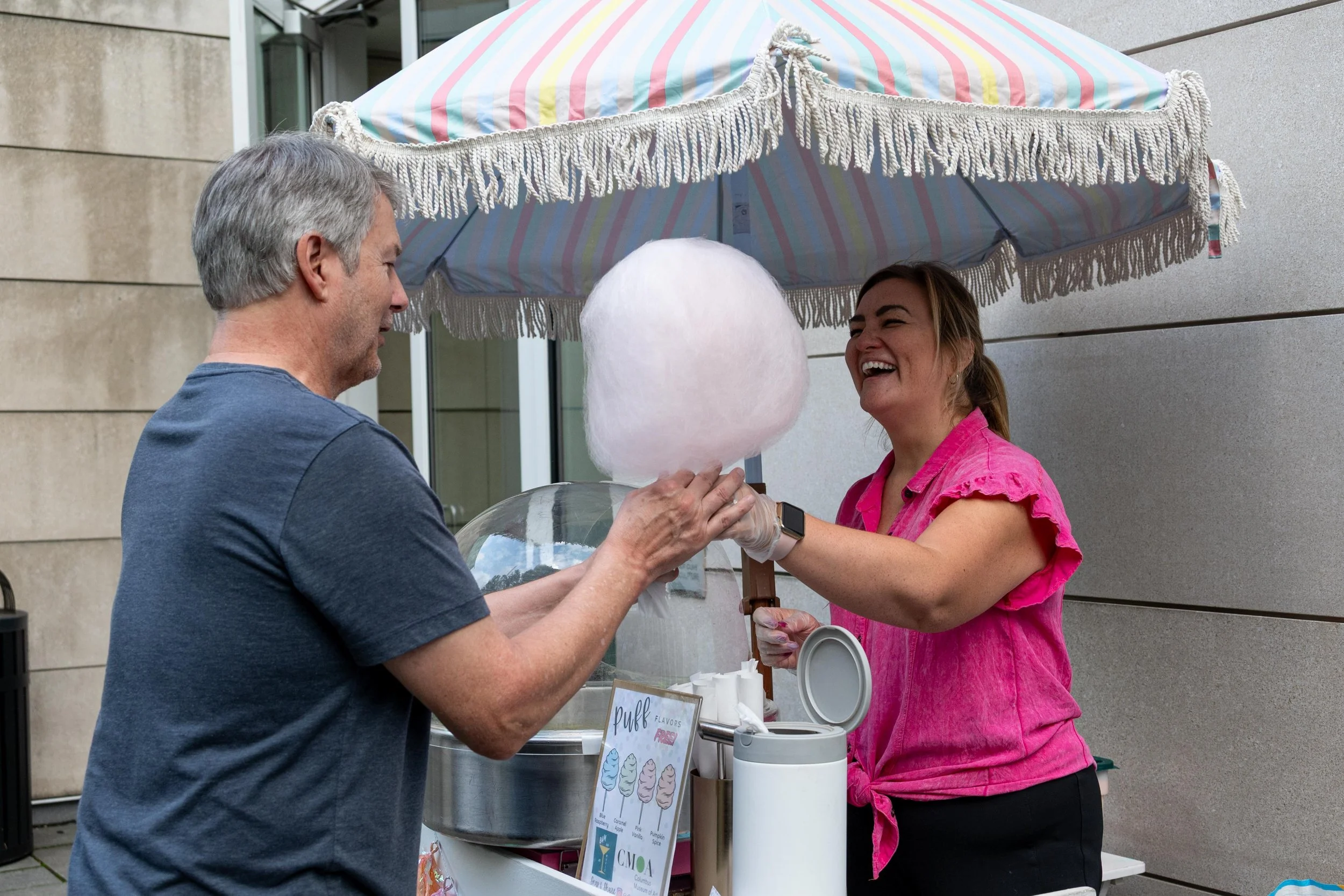 A woman wearing a pink top is smiling and holding a large stick of cotton candy, handing it to a man with gray hair wearing a navy blue shirt. They are outside under a pastel-colored striped umbrella.