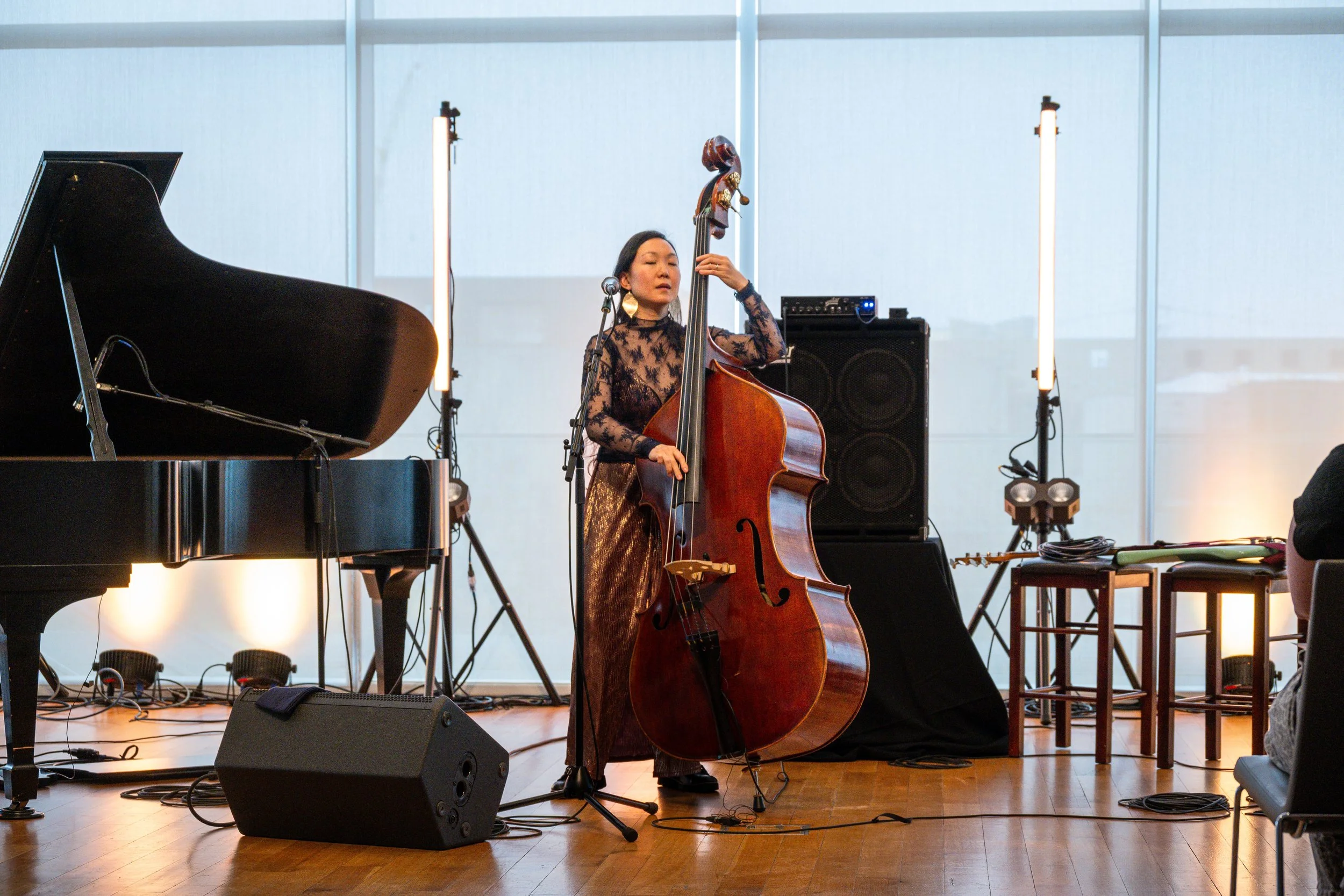 A woman playing a double bass on a stage with a piano nearby, surrounded by musical equipment, in a room with large windows.