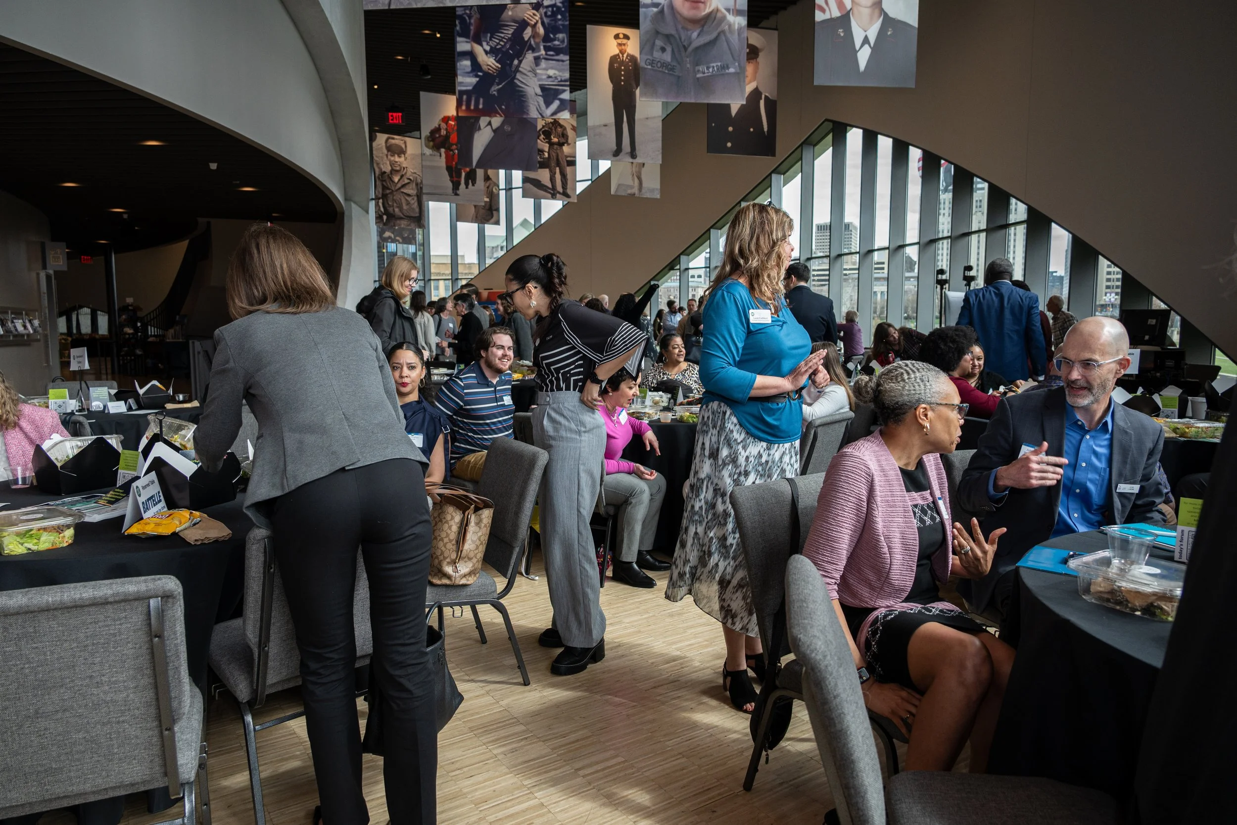 People gathered in a modern, spacious indoor area, some seated at tables with food and drinks, others engaging in conversation or standing. Large portraits of military personnel are hanging from the ceiling, and large windows overlook a cityscape.