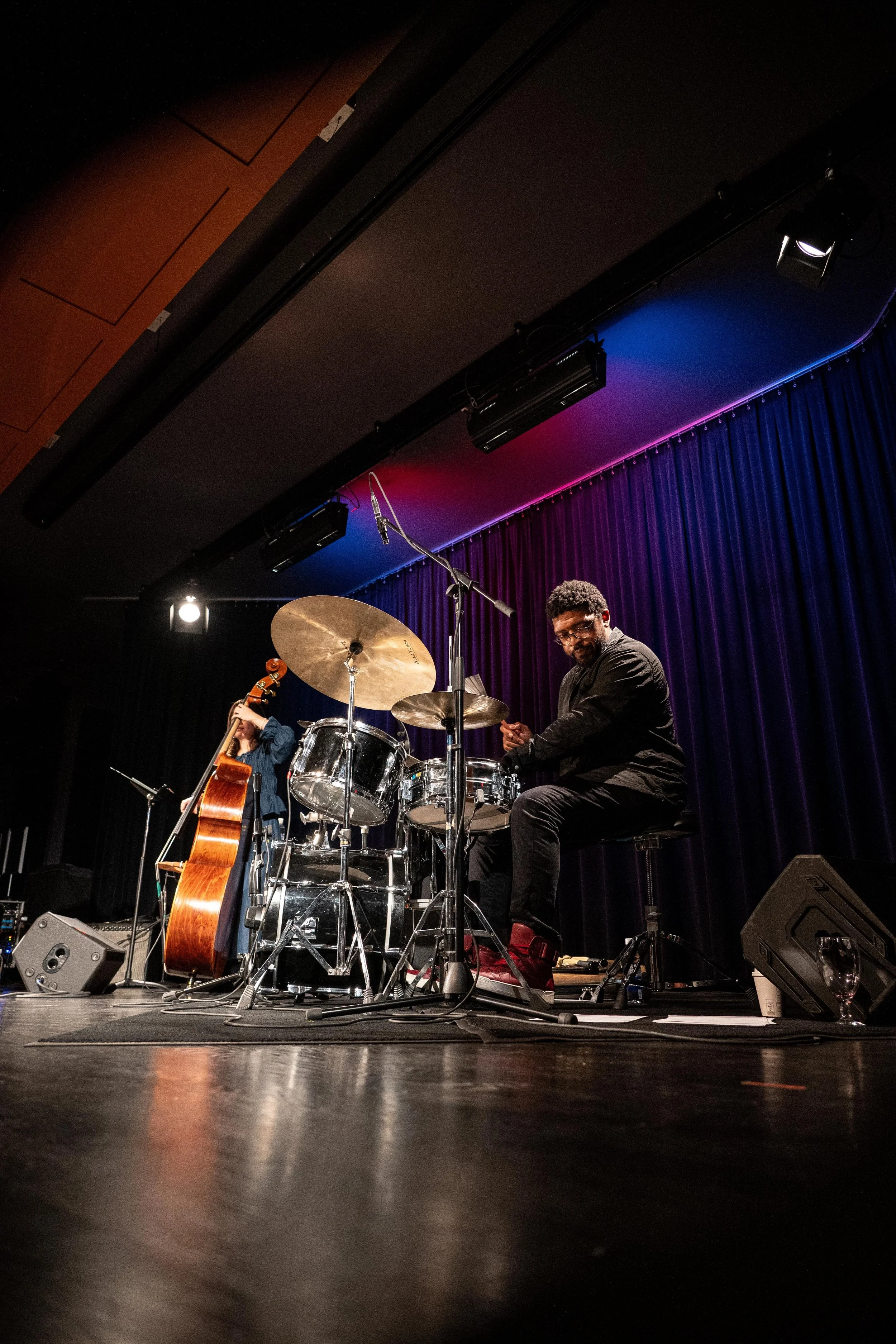 A jazz band performing on stage with a drummer, a cello, and a person playing a double bass. The stage has purple curtains and colored lights.