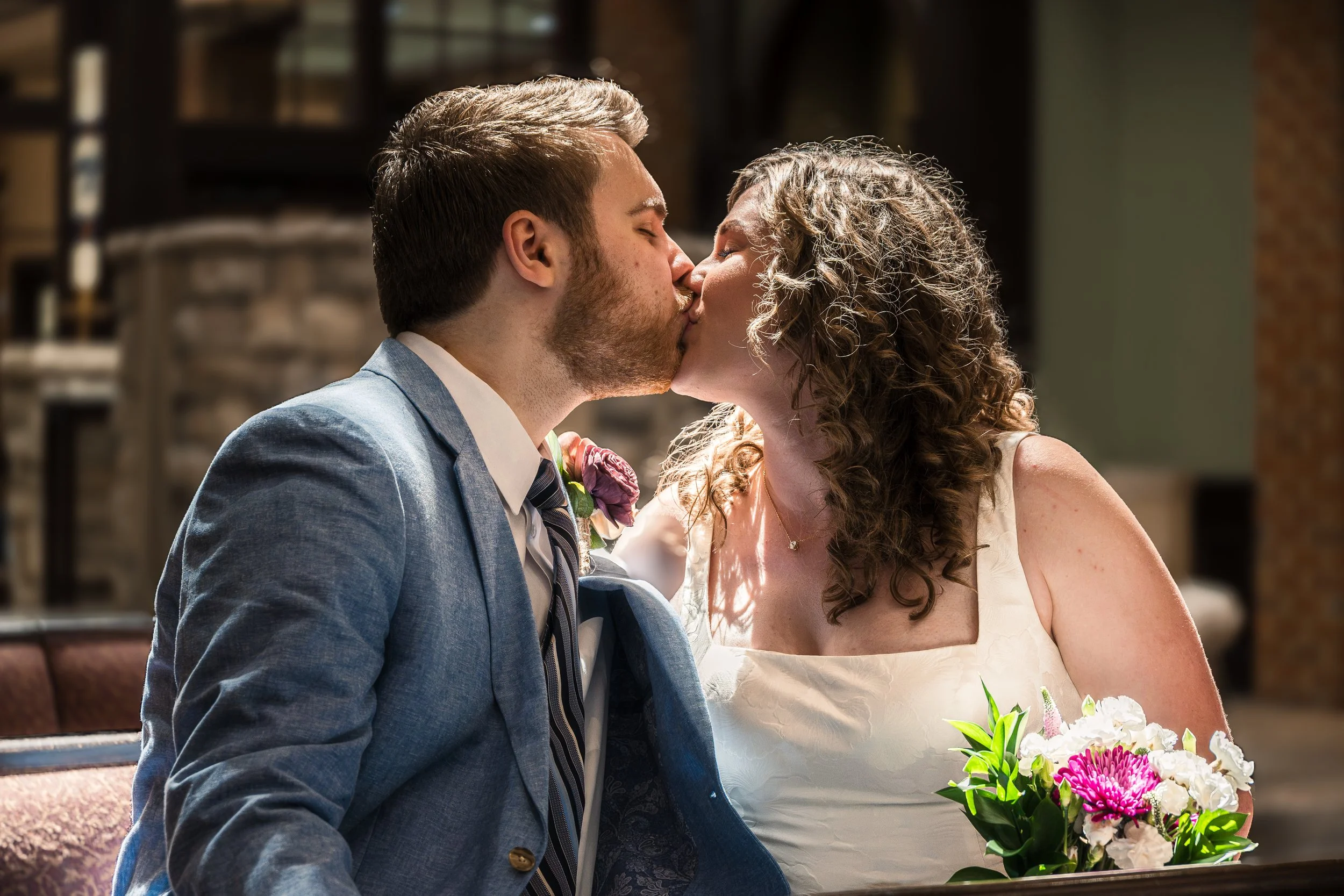 A couple in wedding attire sharing a kiss at their wedding ceremony, with the woman holding a bouquet of colorful flowers.