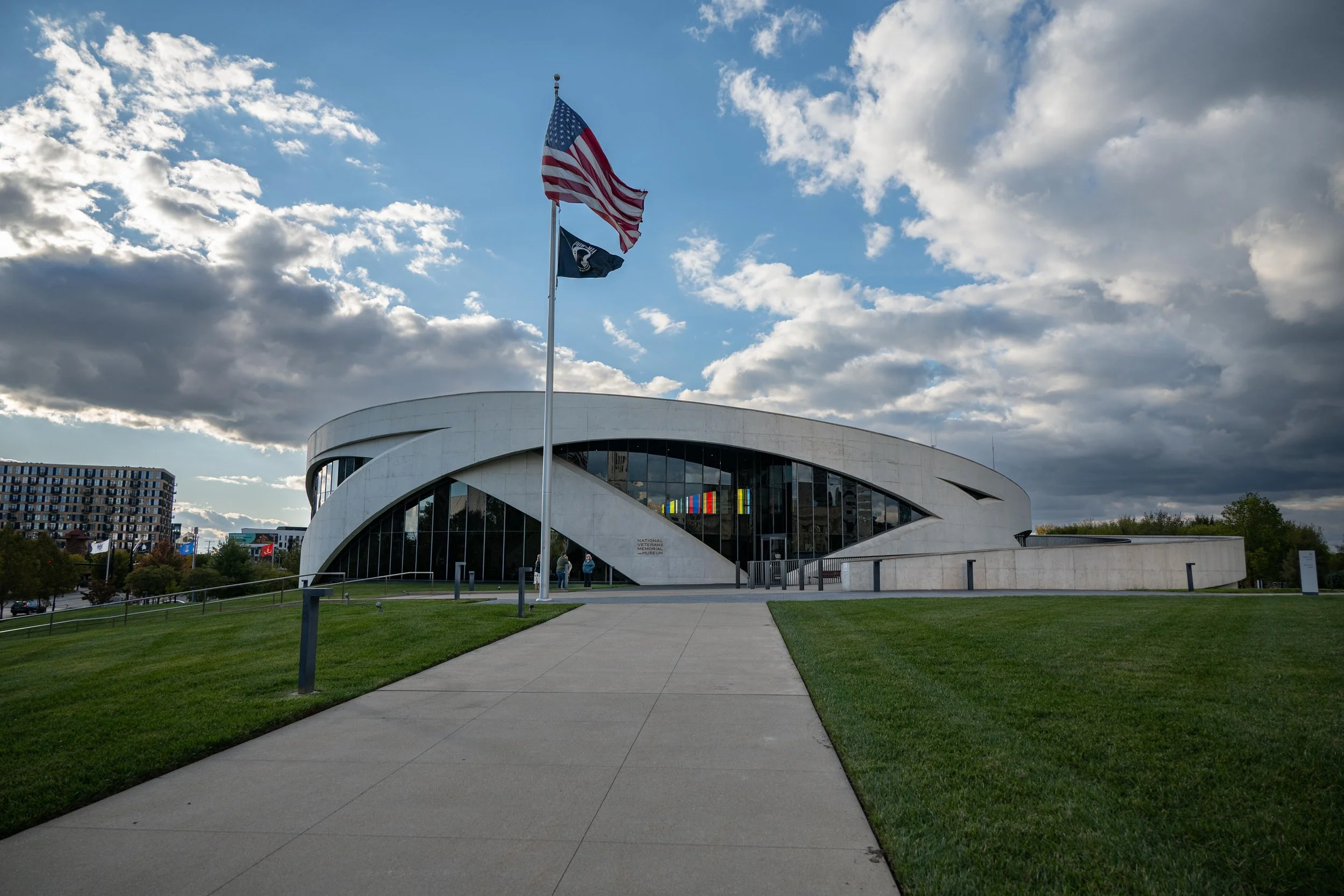 View of the National Veterans Memorial and Museum building with United States flag and a second flag flying in front, under a partly cloudy sky.