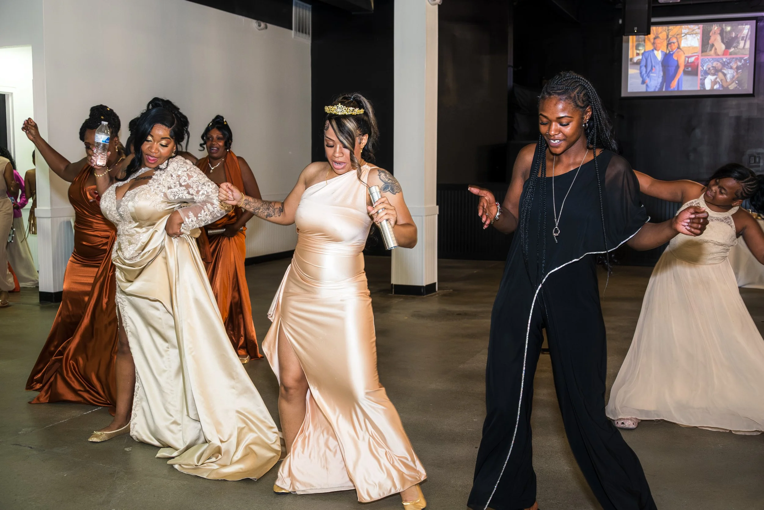 A group of women dancing at a celebration, dressed in elegant gowns, with some wearing jewelry and tiaras. They appear joyful and are enjoying the music, with a screen displaying photos in the background.