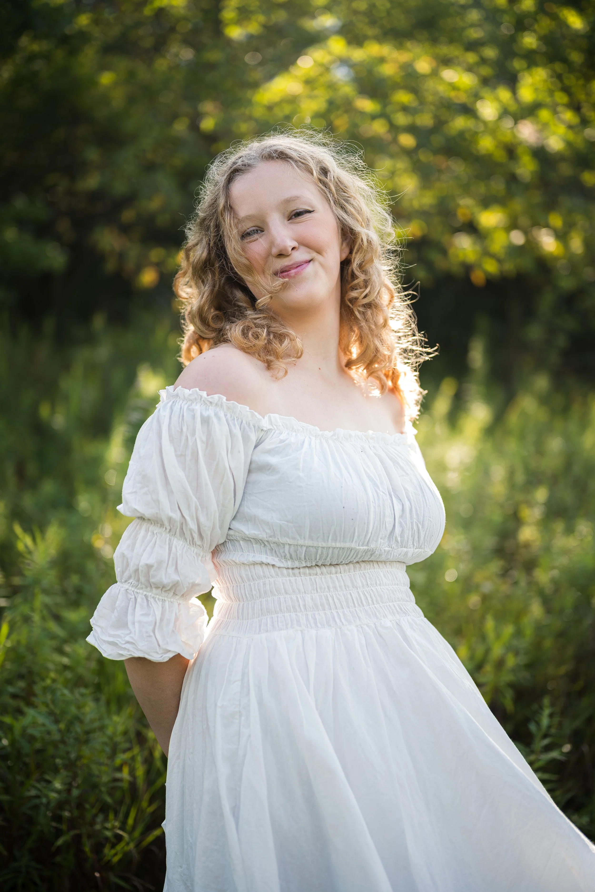 A woman with curly blonde hair wearing a white off-shoulder dress, smiling in a natural outdoor setting with green foliage and sunlight in the background.