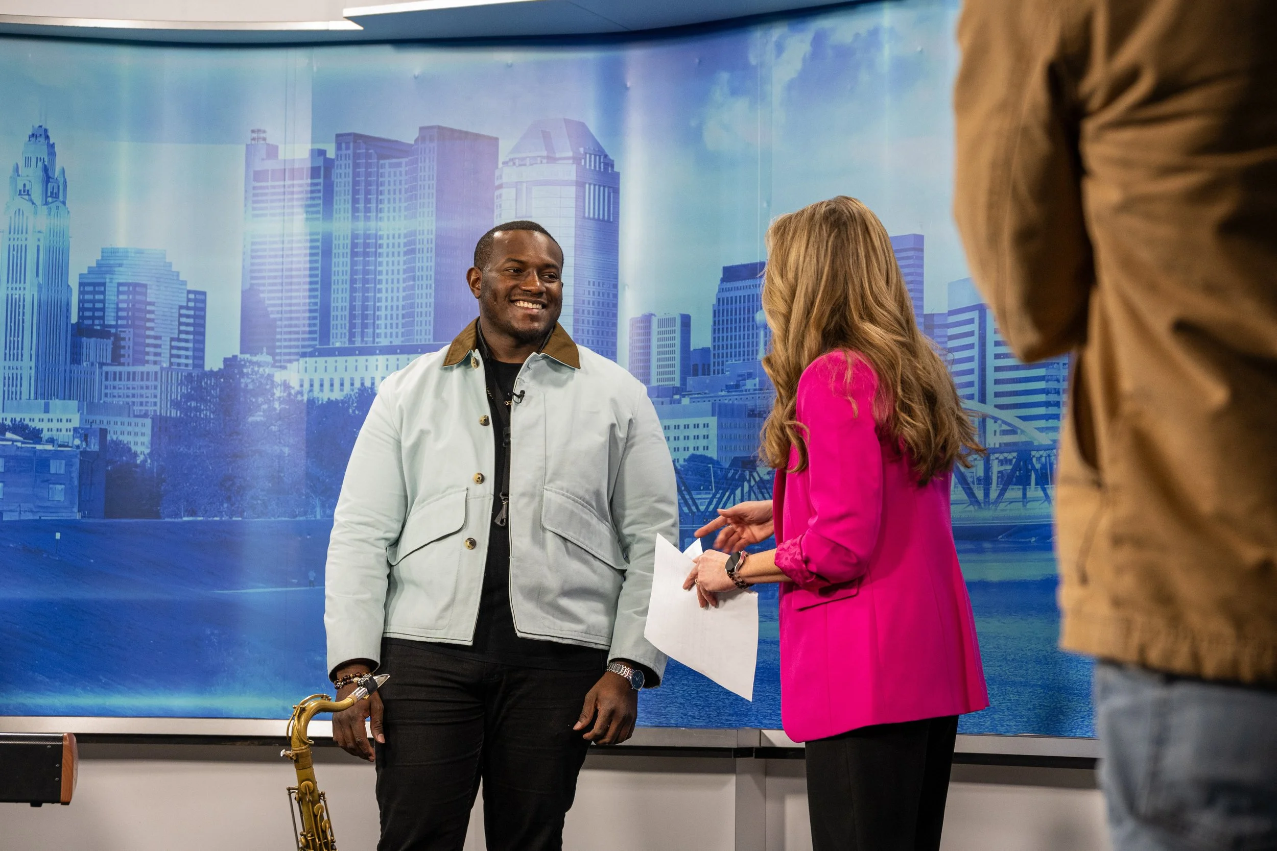 A man with a saxophone standing and smiling while engaged in conversation with a woman in a pink blazer, in front of a blue cityscape backdrop, with a person in a brown jacket partially visible in the foreground.