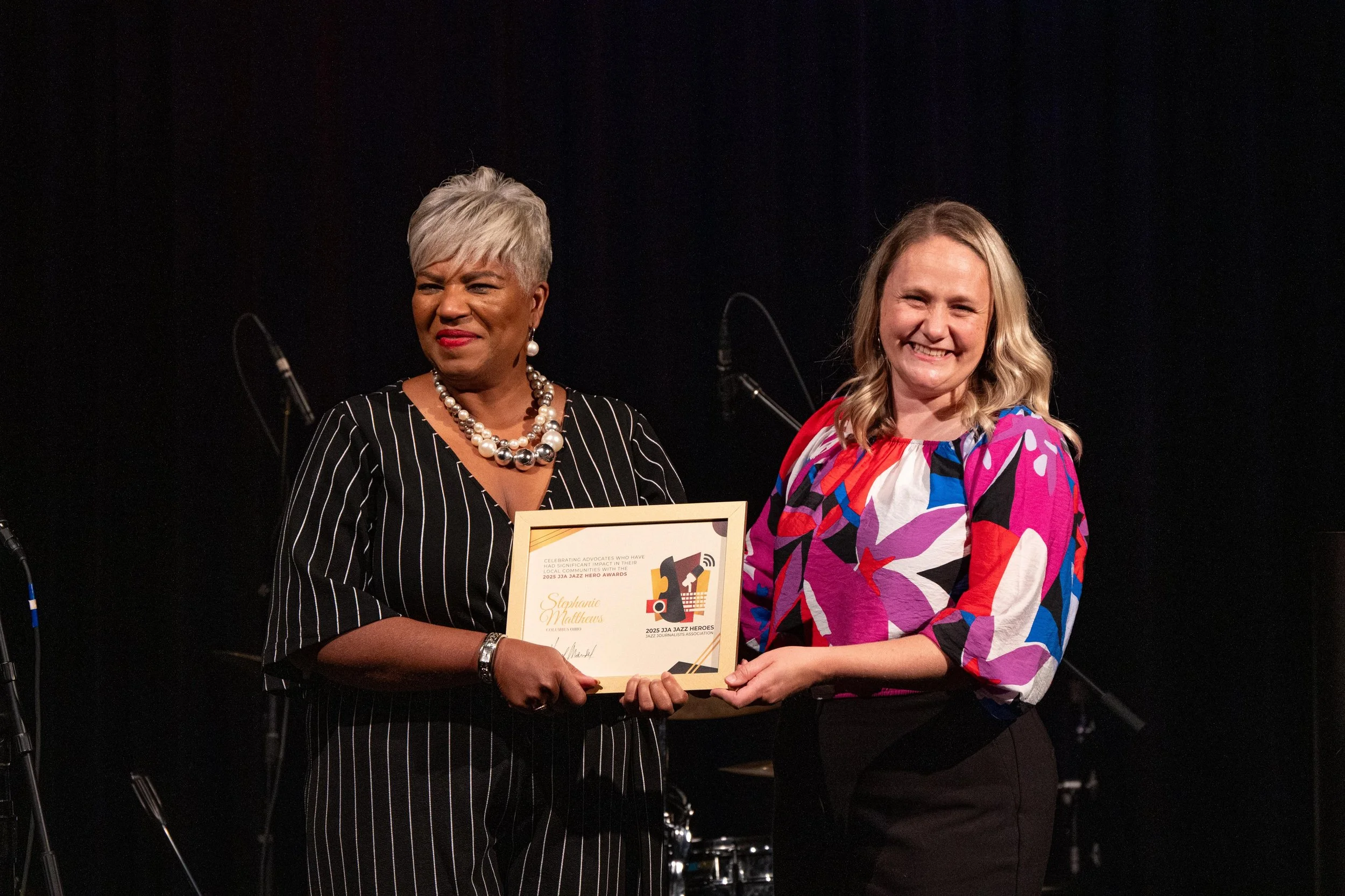 Two women standing on a stage, one woman with short gray hair and a pearl necklace holding a framed award, and the other with long blonde hair wearing a colorful patterned blouse, smiling as they shake hands and pose for a photo against a dark backgr